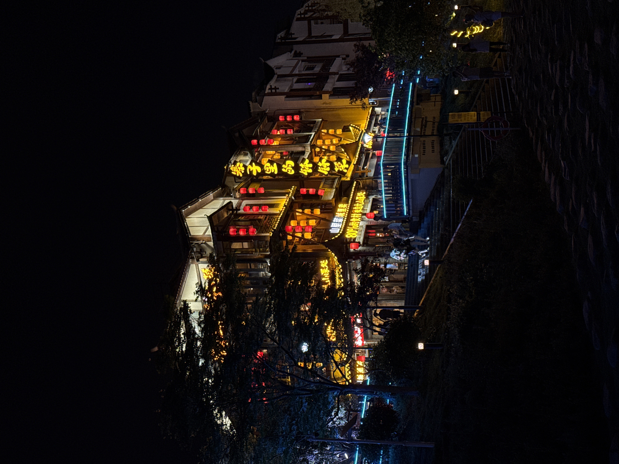 Here's a concise description of the image:
The image shows a building illuminated with yellow and red lights at night. Text, possibly in Chinese, is visible in the yellow lights. A staircase with blue lights runs along the building's side. Trees are visible in the foreground, partially obscuring the view of the building. People are on the staircase. The location appears to be Wulingyuan, China, judging from the architectural style and lighting common in that area.
The OCR text identifies the building as a restaurant ("寨子里的钵体菜" - literally "Bowl Dishes in the Village") that sources its ingredients from the mountains. The text also claims "80% of the ingredients come from the depths of the mountain".