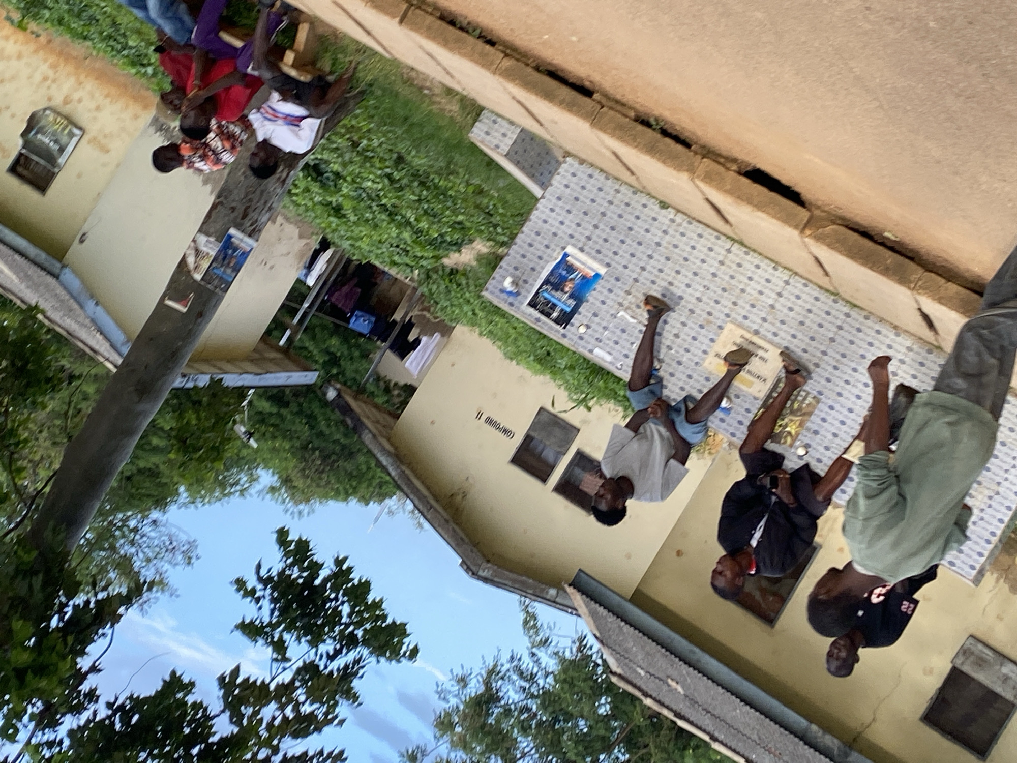 In Rafin Par, Nigeria, a group of young men are gathered outside a building with a pale yellow facade. The building appears to be a residential compound, as indicated by the signage "COMPOUND 11" visible on its exterior. The ground in front of the building is paved with patterned tiles, and a raised concrete ledge runs along the edge of a dirt path.
Several individuals are seated on the tiled area and on a wooden bench. One young man in a light grey t-shirt and blue shorts is seated facing away from the camera. Another man in a black jacket and dark shorts is seated next to him, holding a phone. Further back, three more young men are seated on the bench. One wears a red shirt and purple pants, while another has on a white shirt with red and blue stripes. A third man in a colorful patterned shirt is also on the bench.
In the foreground, another young man in a green t-shirt and grey pants is standing and looking towards the building. To his left, partially visible, is another young man in a black t-shirt. The environment is lush, with green trees and bushes surrounding the building. Laundry items are visible hanging on a line in the background. The overall scene depicts a relaxed outdoor gathering of young men in a residential setting.