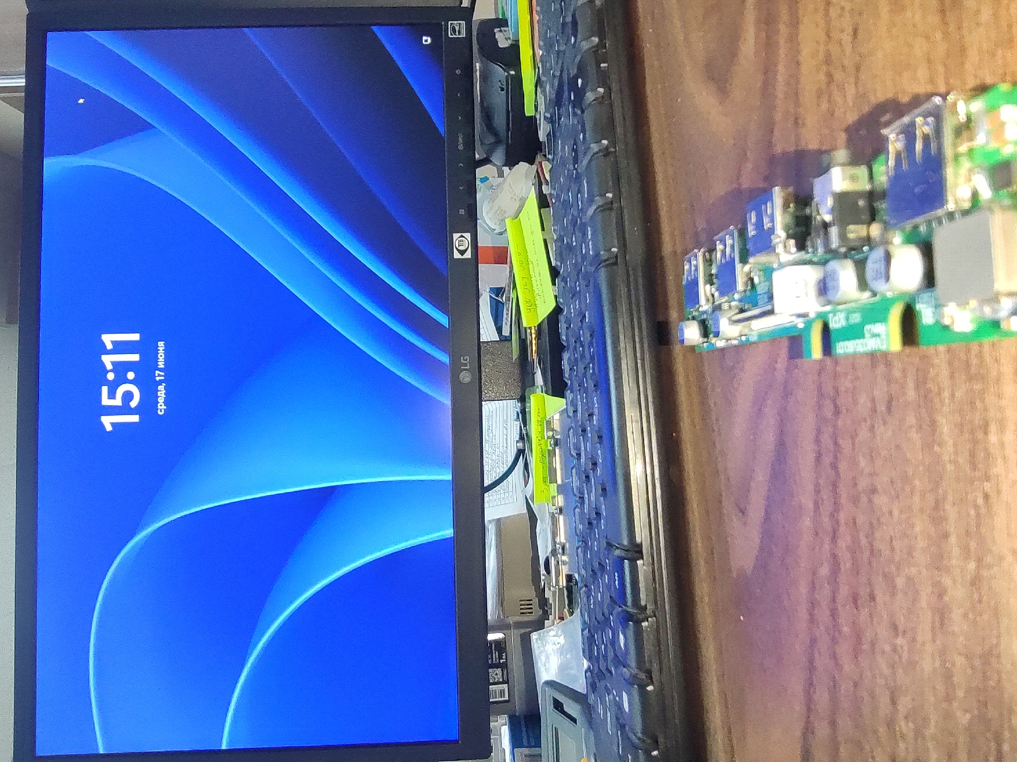 The image shows a computer monitor displaying a time of 15:11 and the date "среда, 17 июня" which translates to "Wednesday, June 17". Below the monitor is a keyboard and several electronic components, possibly a circuit board with USB ports. The scene is set on a wooden surface. There are no people visible in the image. The background suggests an indoor environment, likely an office or workspace.