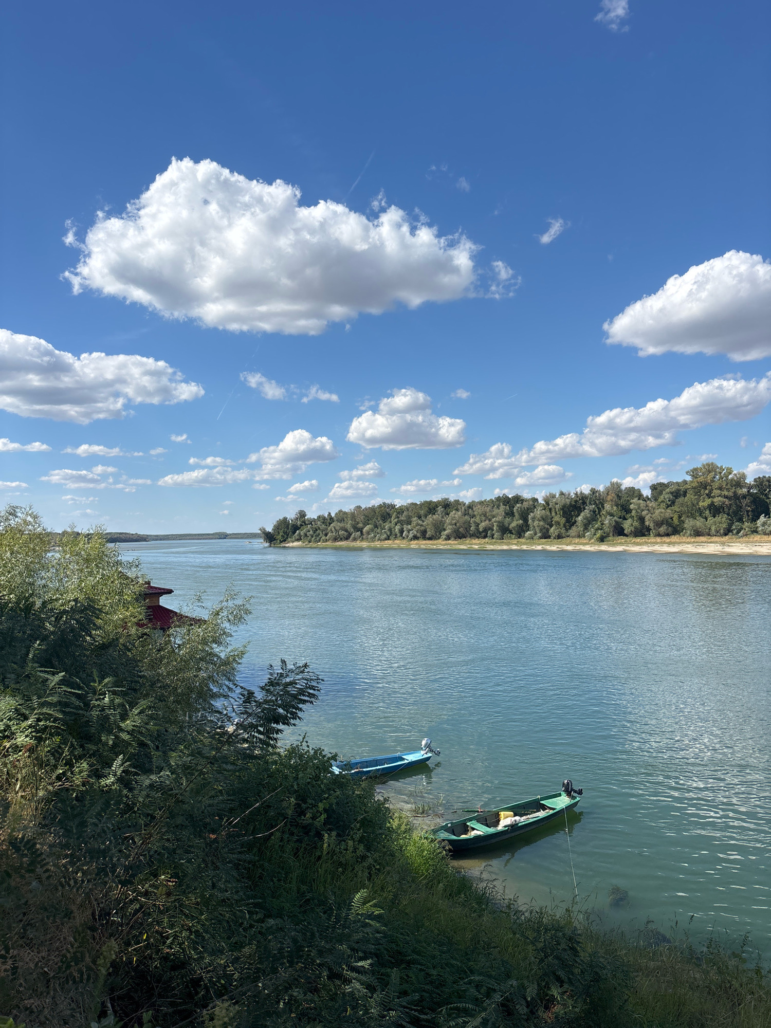 The image shows a wide, tranquil river under a bright blue sky dotted with fluffy white clouds. On the left bank, lush green foliage and trees partially obscure a red-roofed structure. Further out in the river, two small boats are visible. One is a light blue motorboat, and the other is a green motorboat with yellow and white items inside, tied to the bank with a rope. The opposite bank of the river is lined with a dense forest. The scene appears to be a serene natural landscape, likely in the vicinity of Aidemir, Bulgaria, given the context.