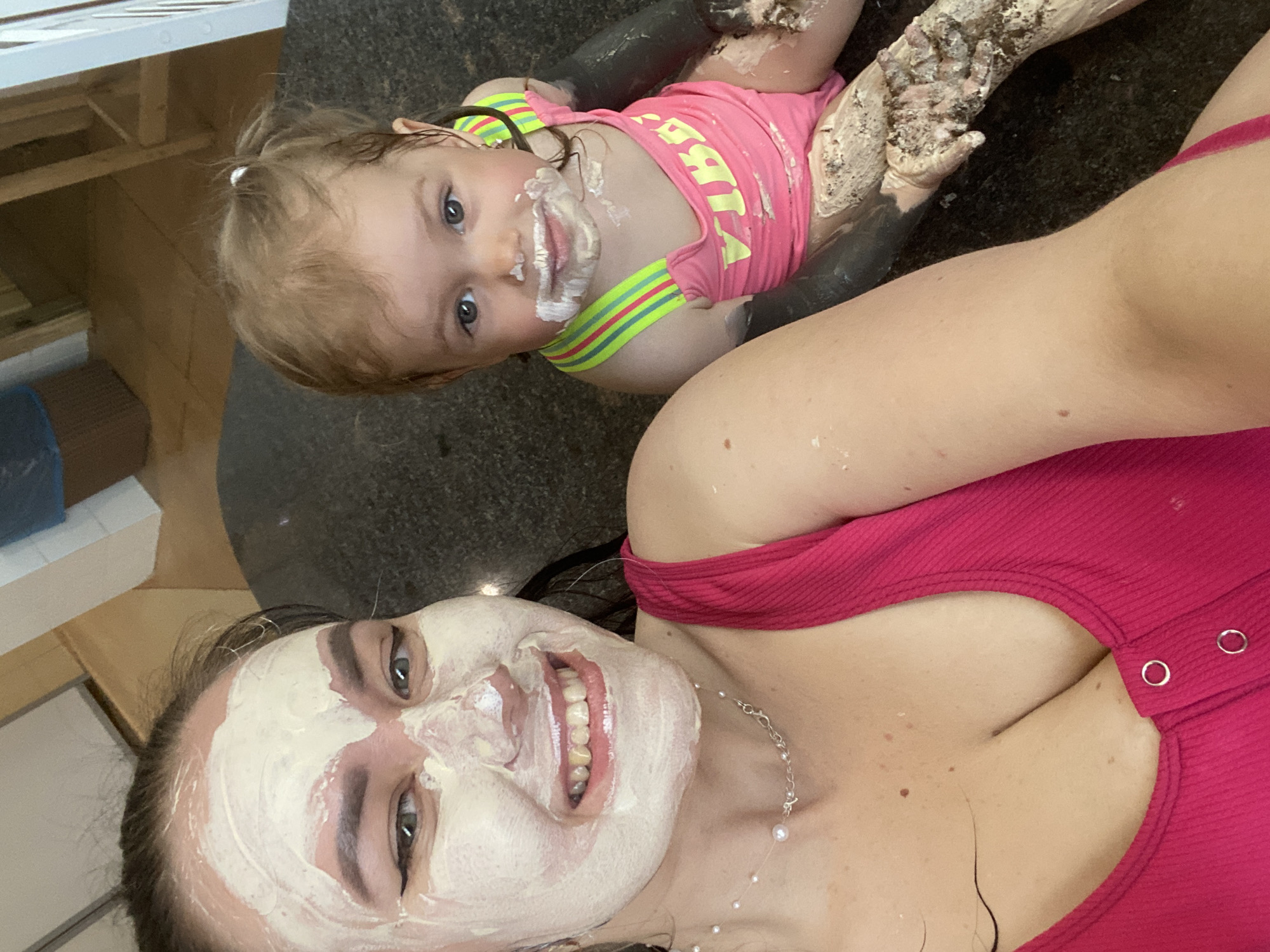 A selfie taken indoors shows a woman and a toddler covered in a white substance, presumably a facial mask. The woman, in the foreground, is wearing a pink top, a necklace, and has a white mask applied to her face. She is smiling broadly, exposing her teeth. Behind her is a toddler wearing a pink top with yellow lettering and colorful trim. The toddler also has white mask residue around their mouth and hands. The background appears to be a kitchen countertop or table. There are other objects visible in the periphery, including what appears to be a white cabinet or shelving unit. The location in Votkinsk, Russia, is not discernible from the image.