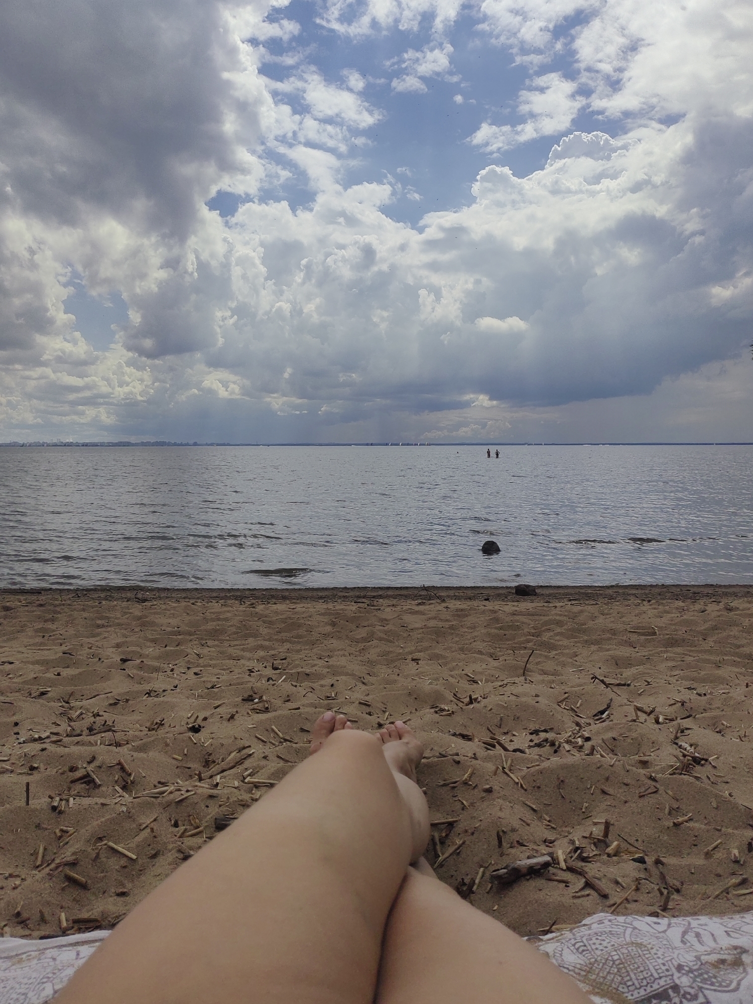 The image depicts a first-person perspective of someone lying on a sandy beach, with their legs crossed. The beach is littered with small pieces of driftwood. Beyond the beach, a large body of water stretches to the horizon, with several sailboats visible in the distance. The sky is partly cloudy, with large cumulus clouds and patches of blue sky. In the far distance, a city skyline is faintly visible. Two people are standing in the water, and a dark, round object, possibly a rock, is visible in the water closer to the shore. The overall scene suggests a summer day at a beach near Saint Petersburg, Russia, possibly along the Gulf of Finland.