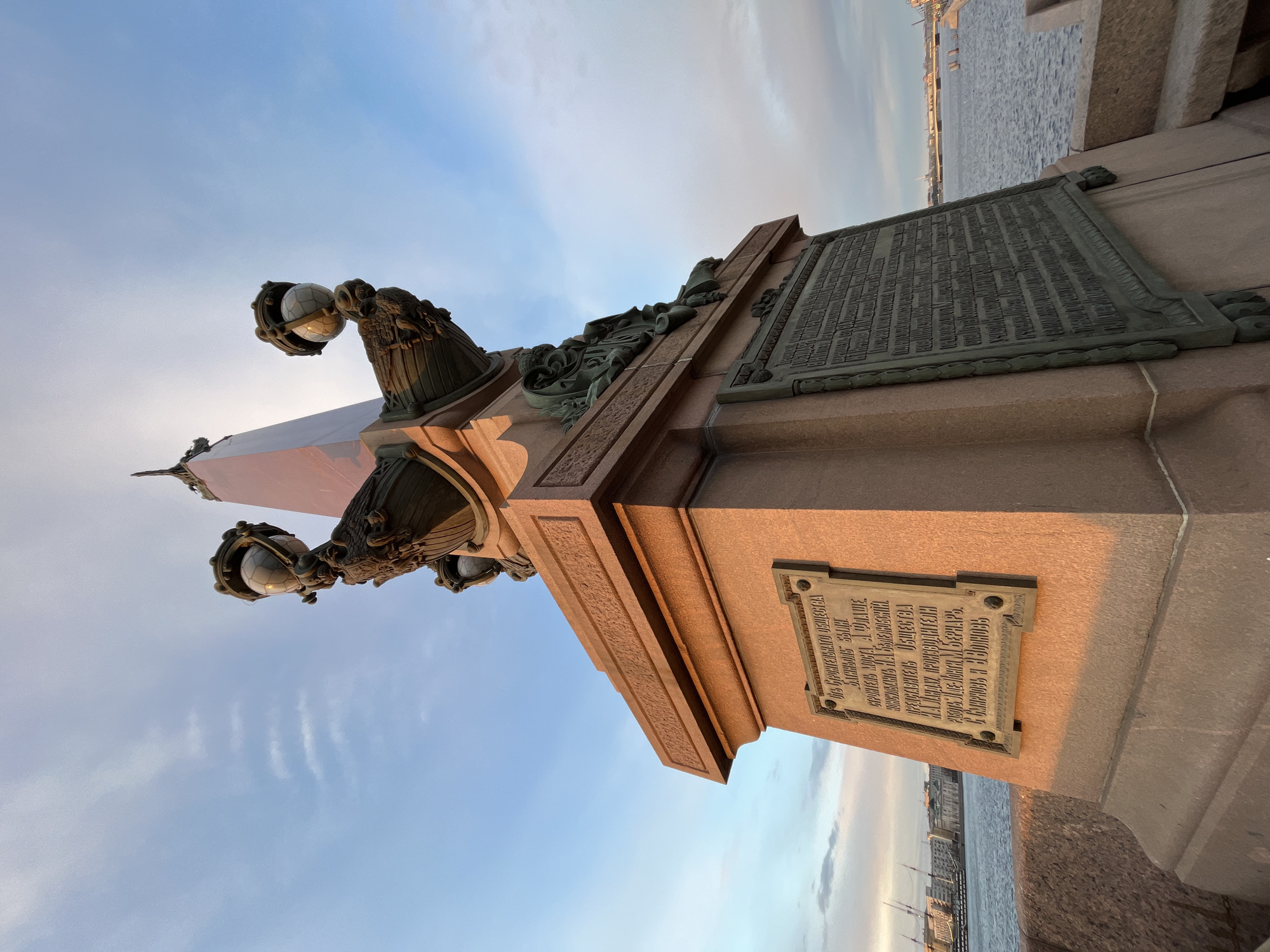 The image shows a lamppost on the Palace Bridge in Saint Petersburg, Russia. The lamppost is made of stone and metal and features decorative elements such as sculptures of mythical creatures. There are inscriptions on plaques affixed to the stone base. In the background, the Neva River is visible, as well as other buildings along the embankment. The sky is visible.
