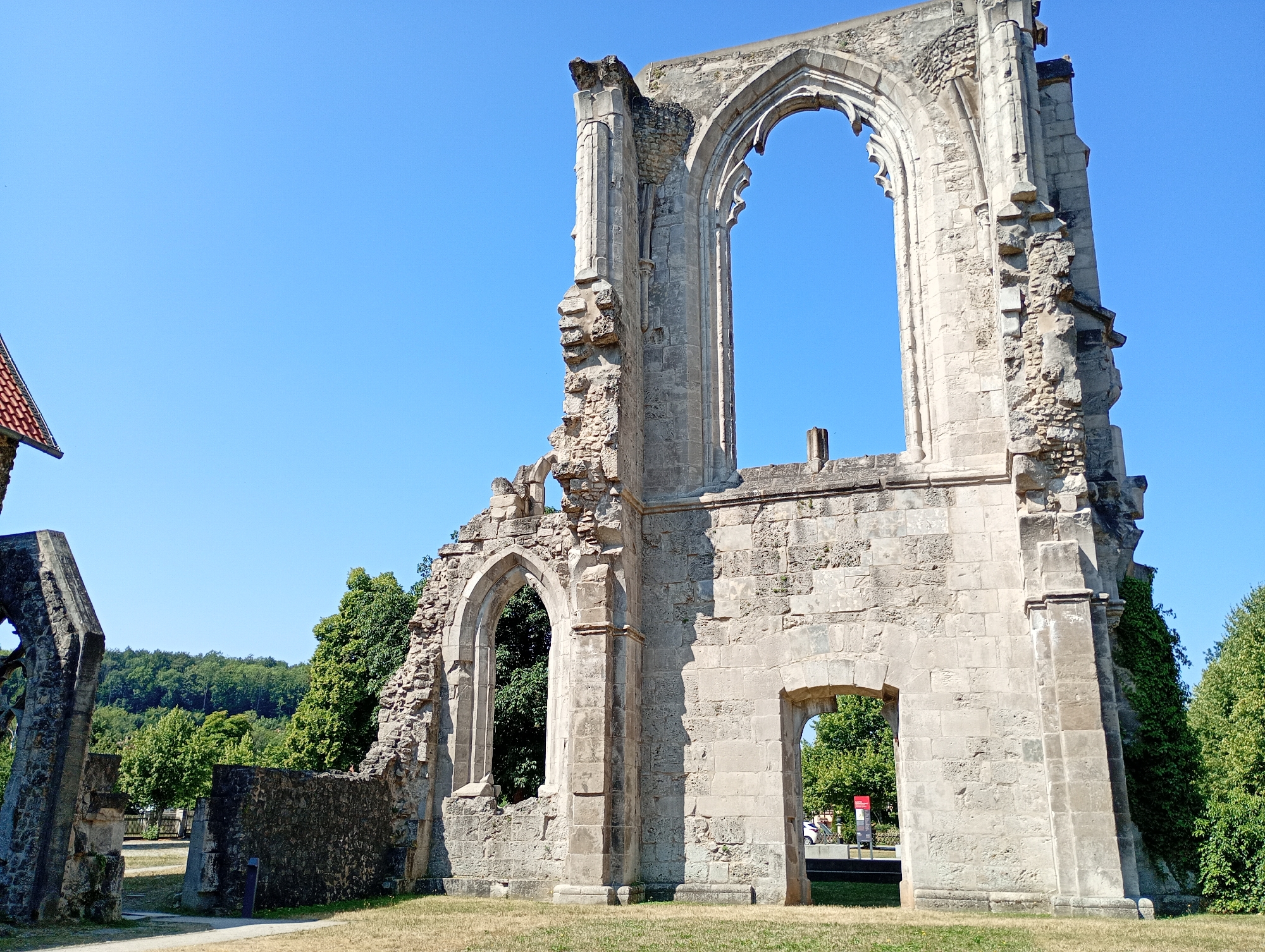 The image depicts the ruins of the former Walkenried Abbey in Walkenried, Germany. The primary subject is a large, stone ruin with prominent arched openings and weathered stone blocks. The sky is a clear, bright blue. In the foreground, dry grass covers the ground. To the left, a portion of a red-tiled roof is visible, suggesting proximity to another structure. Behind the ruins, lush green trees and a forested hillside are present. A narrow path runs through the grass towards the ruins. The overall scene conveys a sense of historical decay and natural overgrowth.