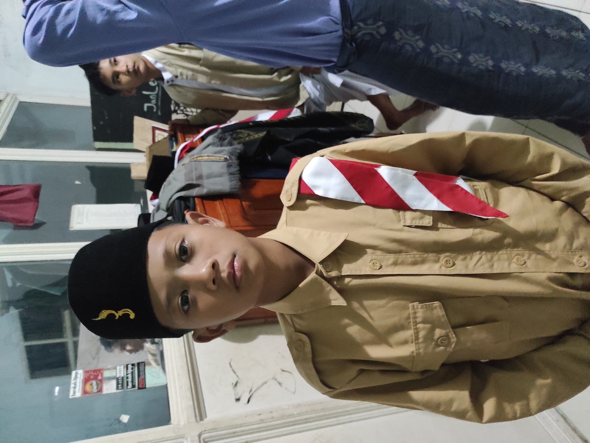 The image depicts a young boy in a scout uniform, wearing a black peci hat with a gold emblem. He is standing in what appears to be a residential setting, possibly indoors or near an entrance. Behind him, another young boy is visible, wearing a light-colored uniform. The background shows a window with stickers, some shelves with items, and a blackboard with writing that includes "JanLe". The lighting suggests it is evening or indoors. The overall scene suggests a gathering or event related to scouting or an educational institution. Given the context of the prompt, this scene is set in the city of Sumedang, Indonesia.