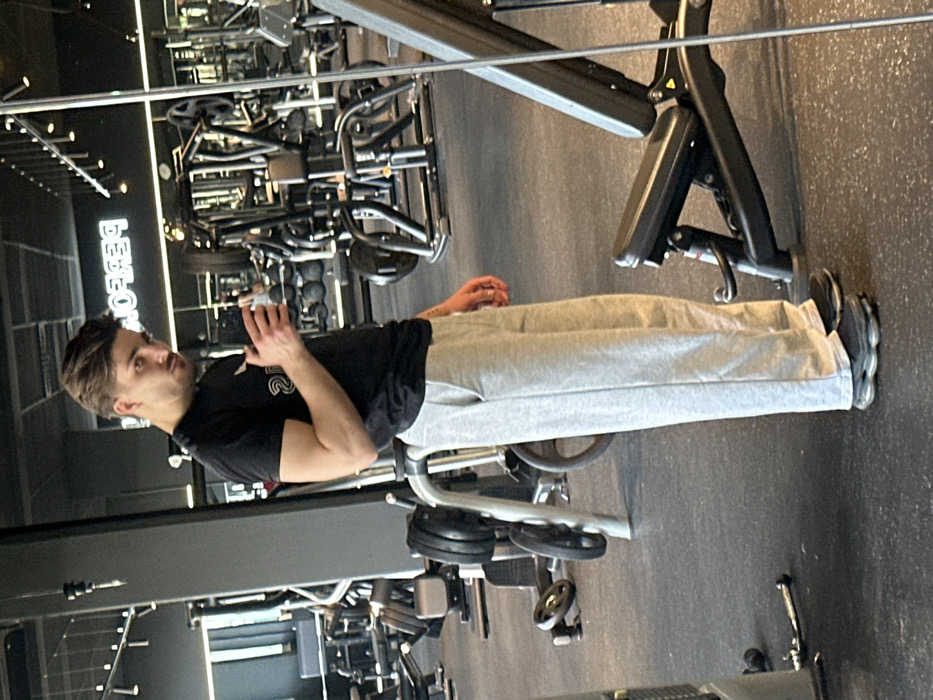 An adult male stands in a gym located in Jestetten, Germany. The individual, with dark, styled hair and facial stubble, wears a black short-sleeved t-shirt, light grey sweatpants, and dark athletic shoes with segmented soles. He holds a black smartphone in his raised right hand, consistent with the act of capturing a self-portrait in a mirror. His gaze is directed towards the camera's perspective. The gym environment features dark rubberized flooring and dark walls. In the background, reflected in a large mirror, are various pieces of exercise equipment including weight racks, cable machines, and benches. A reversed, illuminated sign displaying "PERFORMA" is visible on the distant wall's reflection. A black adjustable weight bench is partially present in the lower right foreground.