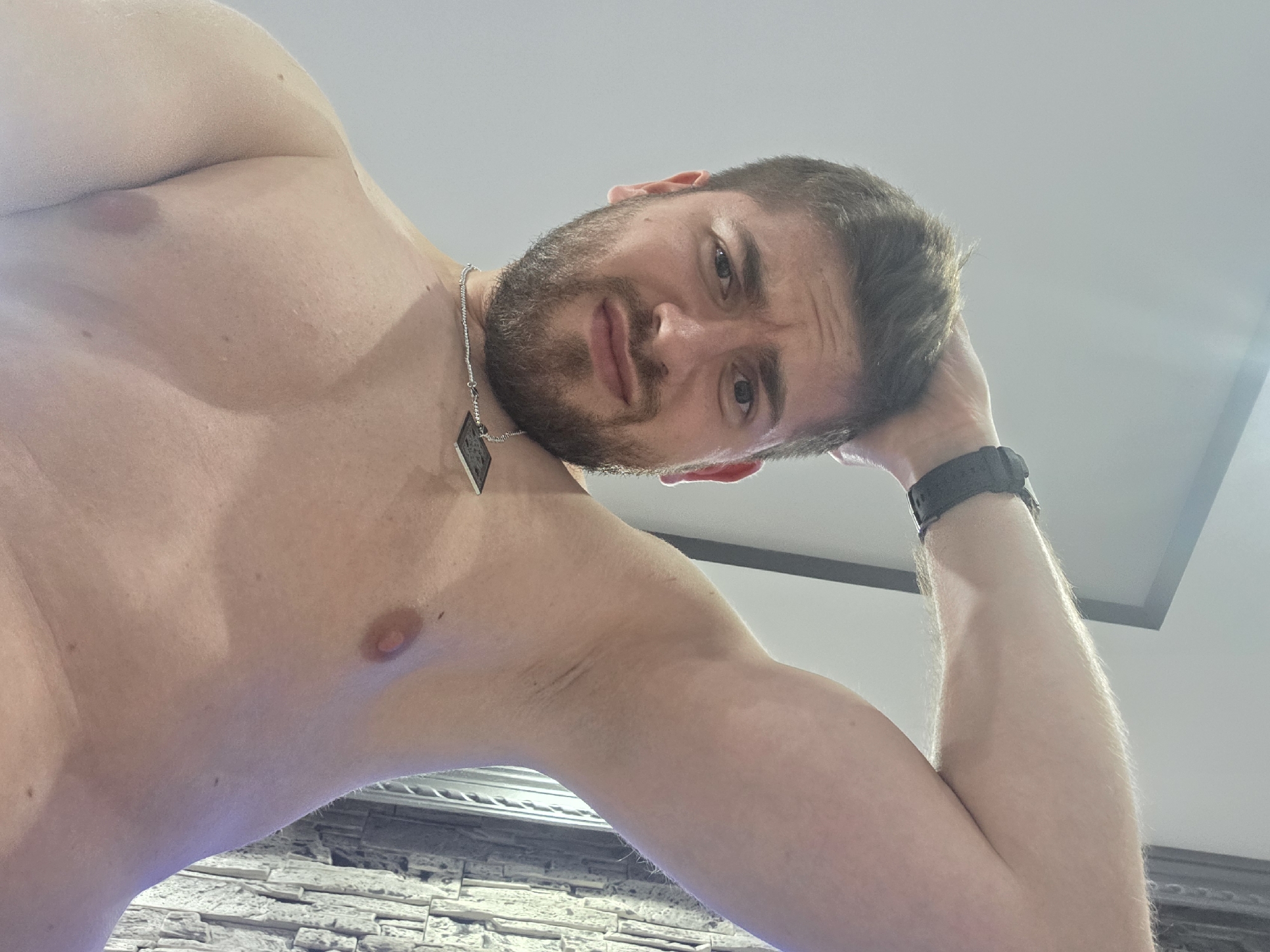 A shirtless man with a beard and short brown hair looks down at the camera. He wears a silver chain necklace with a dark pendant and a black wristwatch. His right arm is raised, with his hand near his head. The background includes a white ceiling and a stone-like textured wall. There is no evidence to suggest that the image was taken in Yagodovo, Bulgaria.