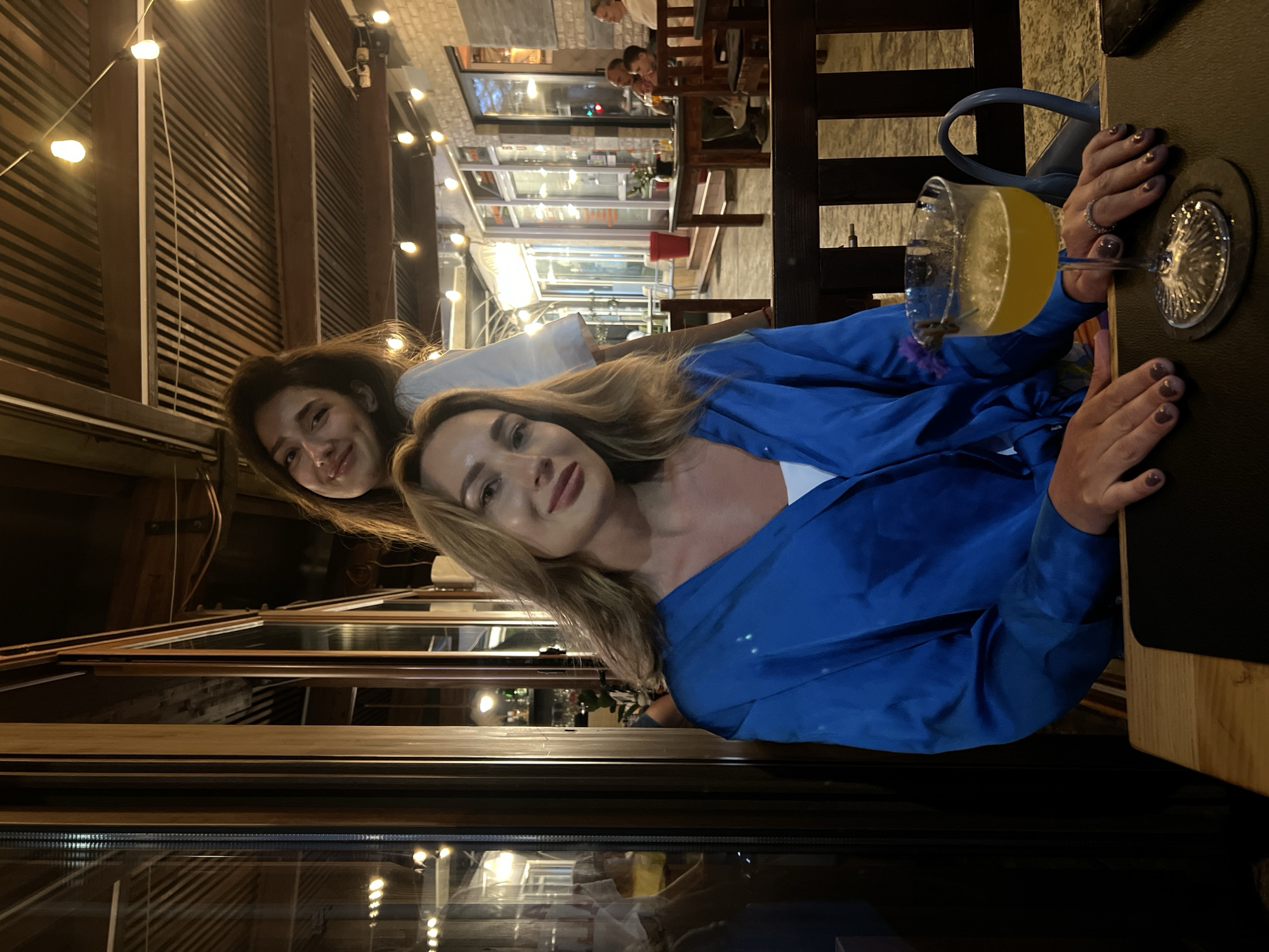 The image depicts two women within a covered outdoor or semi-enclosed restaurant patio in Bar, Montenegro, at night. The setting is dimly lit by numerous string lights strung across a slatted wooden ceiling with exposed beams.
In the foreground, a blonde woman with long hair, wearing a royal blue satin-like long-sleeved shirt over a white top, is seated at a wooden table. Her hands, with dark manicured nails and a ring on her right ring finger, rest on a dark placemat next to a stemmed glass containing a yellow liquid garnished with a small purple flower. A dark smartphone is also visible on the table to her right. She maintains direct eye contact with the camera with a slight smile.
Behind her, another woman with dark hair and a white top stands, leaning forward and looking over the first woman's left shoulder, smiling towards the camera.
The background reveals further seating areas with dark wooden tables and benches. On the right, indistinct figures, including an adult male and a child, are seated at a table. The far background shows buildings with large glass windows and light-colored brick facades. On the left, large glass panel windows with dark wooden frames reflect interior lights. The ground outside appears to be paved.