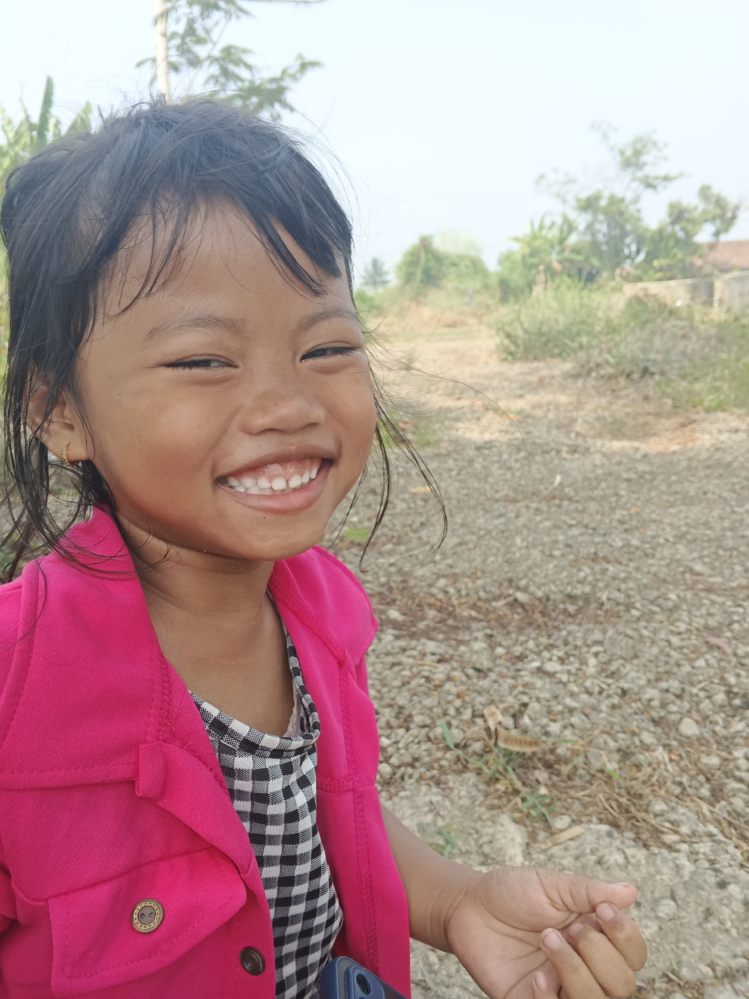 A young girl with dark hair and a bright smile is the subject of this close-up photograph, taken outdoors. She is wearing a bright pink jacket over a black and white checkered shirt. Her right eye is partially closed, and her mouth is open, revealing her teeth. A small gold earring is visible in her left ear. The background suggests a rural or suburban setting, with a dirt path, gravel, and sparse vegetation. In the distance, there are trees and a building with a reddish roof. The sky is bright and hazy, indicating it is daytime. The image appears to have been taken in Sukabumi, Indonesia.