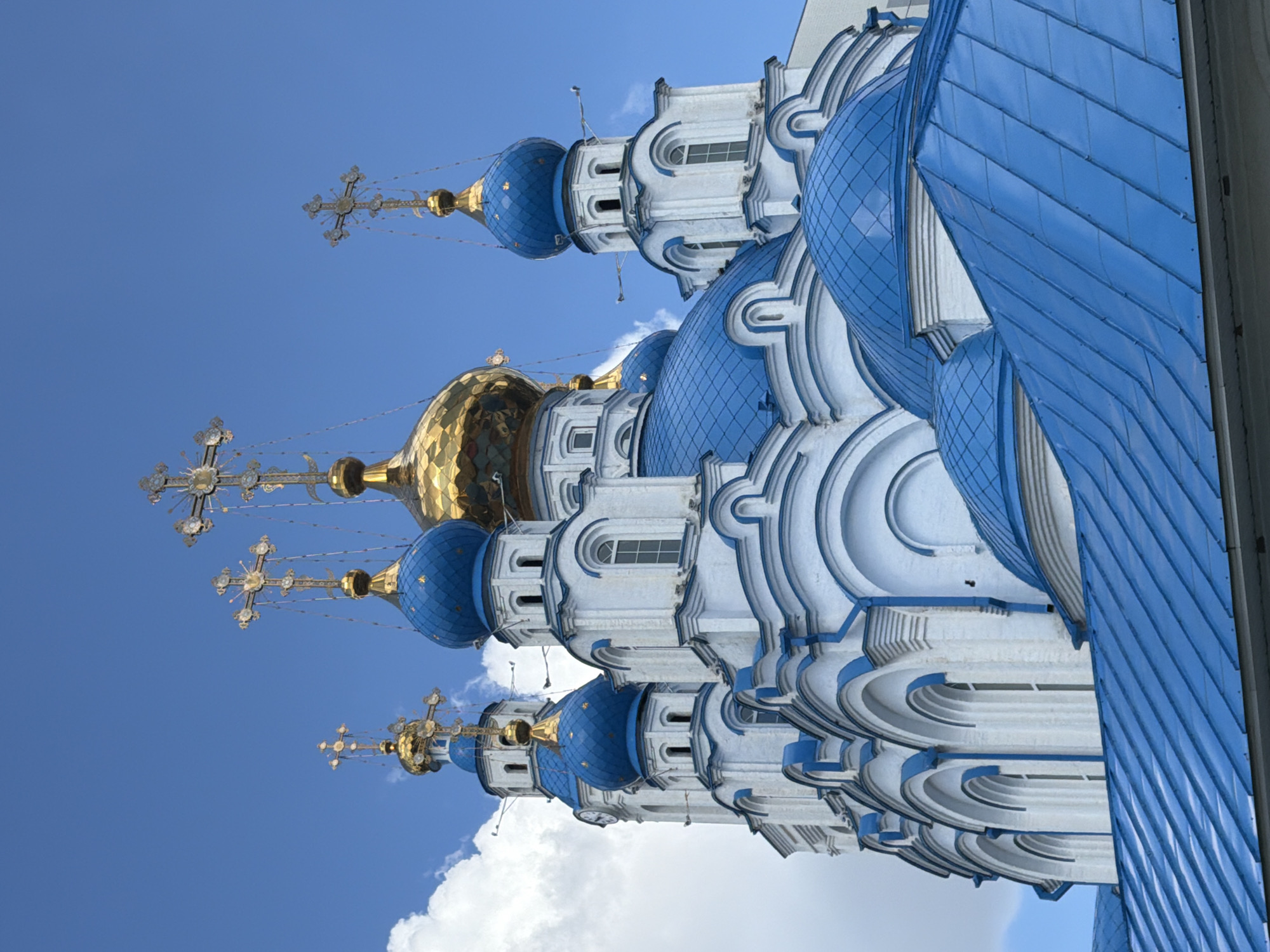 A low-angle shot captures the upper portion of a white Orthodox church with multiple blue domes topped with golden crosses, set against a bright blue sky with scattered white clouds. The church features white walls with blue decorative trim along the rooflines and window frames. Several domed structures are visible, with the largest being a golden dome towards the center of the frame. Smaller blue domes, some with ornate golden crosses, are distributed around the main structure. The foreground is dominated by a blue-tiled roof of another part of the church, angled upwards towards the main building. The architectural style is characteristic of Russian Orthodox churches.