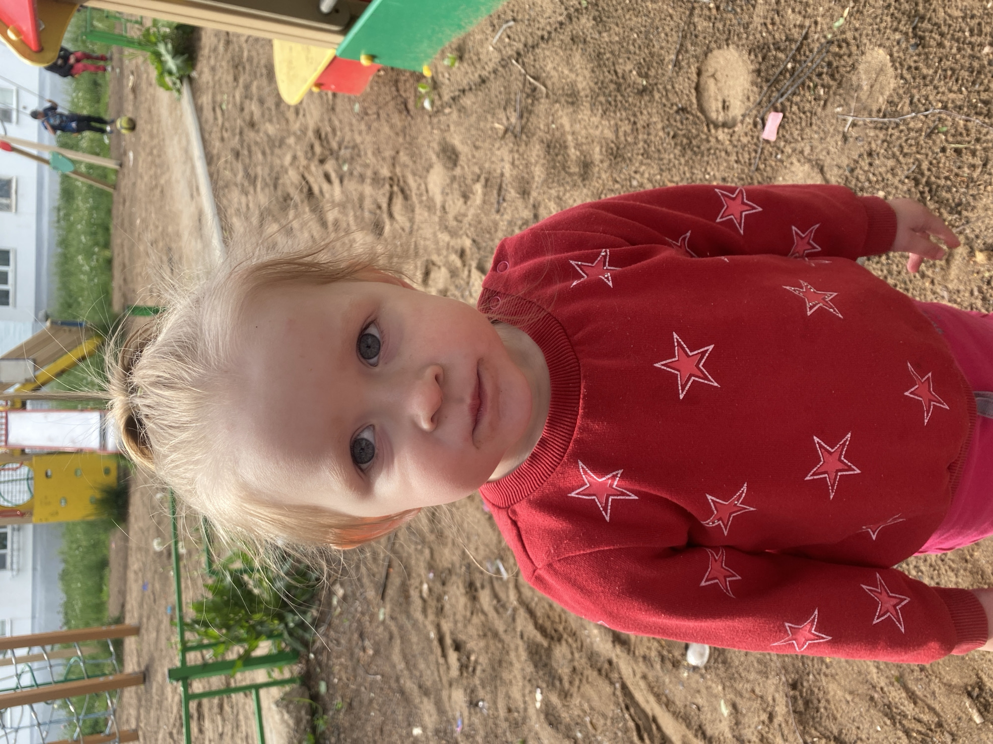 A toddler stands on a sand-covered playground in Chaikovsky, Russia. The child has light blonde hair pulled into a small ponytail on top of their head, and greyish-blue eyes. They wear a red long-sleeved shirt with white star patterns and pink pants. In the background is playground equipment painted in yellow, green, and red. Two figures are playing a game with a ball in the distance. The sky is not visible.