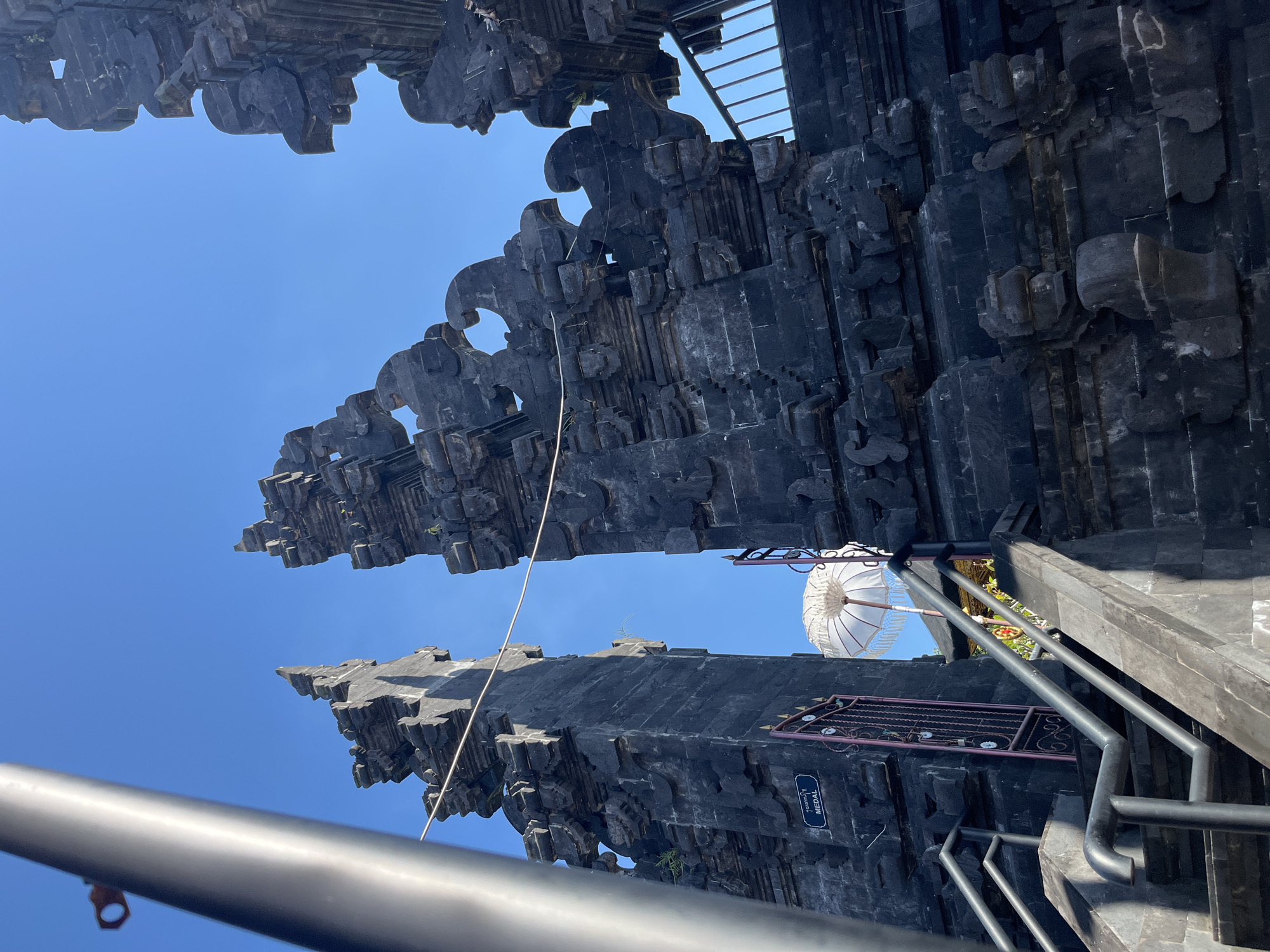 The image shows a low-angle view of the entrance to a Balinese temple, specifically the Pura Taman Beji in Mendoyo, Indonesia. The dominant features are the intricately carved, dark stone gates, characteristic of Balinese architecture. These gates are tall and imposing, with multiple tiers of decorative elements and sharp, pointed finials reaching towards a clear blue sky.
In the foreground, a grey metal railing lines a stone staircase leading up to the entrance. To the left of the stairs, a dark metal gate with ornate scrollwork is partially visible, and above it, a sign with the text "MEDAL" is affixed to the stone wall.
Beyond the left gate, a white ceremonial umbrella with decorative fringe is prominently displayed, along with hints of greenery and what appears to be a shrine area with colorful offerings or decorations. A light-colored wire stretches horizontally across the frame, connecting the two parts of the gate structure. The overall scene is bathed in bright sunlight, casting shadows that emphasize the texture and depth of the stone carvings. The absence of people in the immediate foreground suggests the photo was taken during a quiet moment.