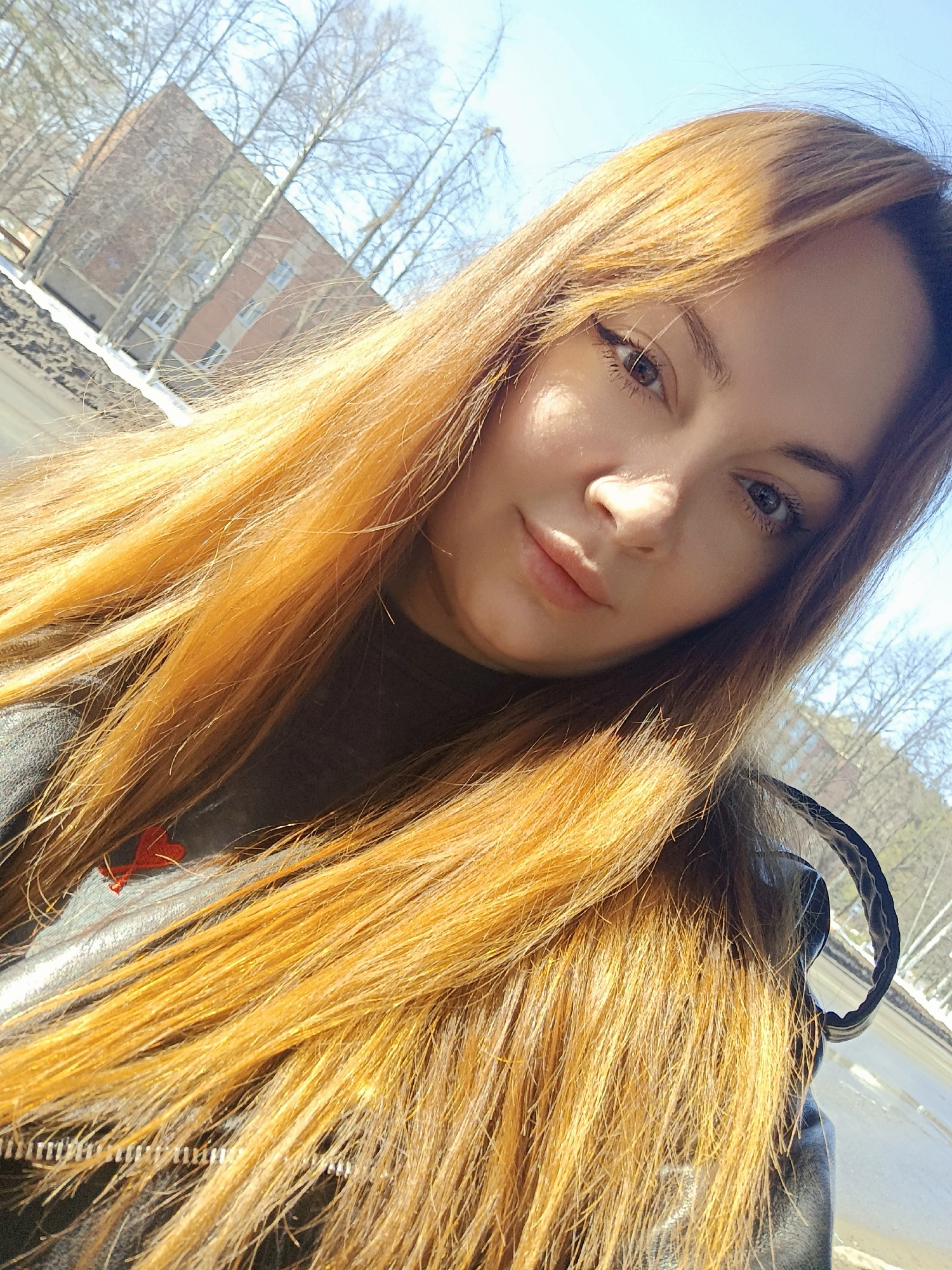 A female individual, appearing to be in her twenties or thirties, is captured in a close-up, chest-up selfie on a clear, sunny day. She has long, straight, reddish-blonde hair cascading over her left shoulder and partially obscuring a dark, high-collared garment and a dark outer jacket, possibly leather. A small, red, heart-shaped emblem is visible on her inner garment near her left shoulder. Her face is turned slightly to the viewer, with direct eye contact, light-colored eyes accentuated by eyeliner, and full lips. Strong sunlight illuminates the right side of her face and her hair.
The background depicts an outdoor environment under a clear blue sky, indicative of late winter or early spring in Chaikovsky, Russia. Numerous deciduous trees with bare branches are visible. In the mid-distance, a multi-story brick residential building with visible windows is present. Further beyond, a wide expanse of what appears to be a partially frozen or snow-lined body of water, likely the Votkinsk Reservoir or Kama River, is discernible, with snow on its banks or edges. The precise location within Chaikovsky, Russia, is not identifiable by specific landmarks beyond a general urban or semi-urban setting overlooking residential architecture and a major waterway, viewed from an elevated perspective such as a balcony or window.