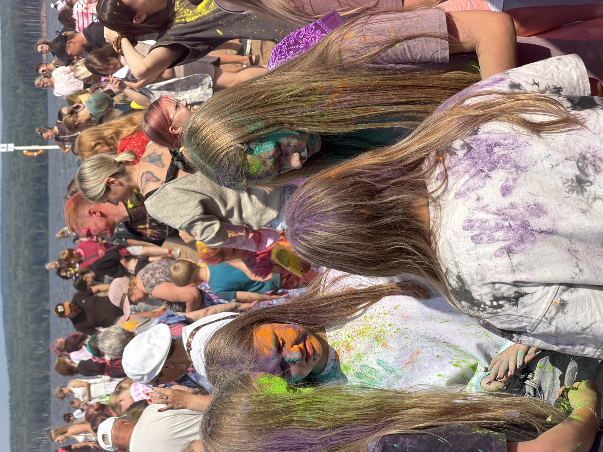 The image depicts a crowd of people celebrating a festival, likely the Holi festival, given the abundance of colored powder on their faces and clothing. The setting appears to be a lakeside or riverbank, with trees and a body of water visible in the background. The sky is clear and sunny, indicating warm weather.
In the foreground, several young girls are prominently featured, their faces and hair adorned with vibrant Holi powder in shades of green, orange, and purple. They are dressed in casual clothing, and some of them are splattered with the colored powder. Their expressions range from playful to contemplative.
Behind the girls, a diverse group of people of various ages are gathered. Some are smiling and interacting, while others appear to be observing the festivities. Many individuals have traces of colored powder on them, suggesting active participation in the event. The atmosphere seems lively and festive.
The environment includes a shoreline with some vegetation and a distant treeline. The water body is calm and reflects the sunlight. The overall scene is one of a communal outdoor celebration. The presence of people dressed in a variety of clothing styles, from casual to more traditional, indicates a broad range of attendees. The organization of the event is not explicitly shown, but the gathering of a large number of people suggests a planned festival or event.
The provided information states this location is in the city of Chaikovsky, Russia. This context suggests that the celebration is taking place on the Kama River embankment, which is a prominent feature of Chaikovsky and a common location for public gatherings and festivals.