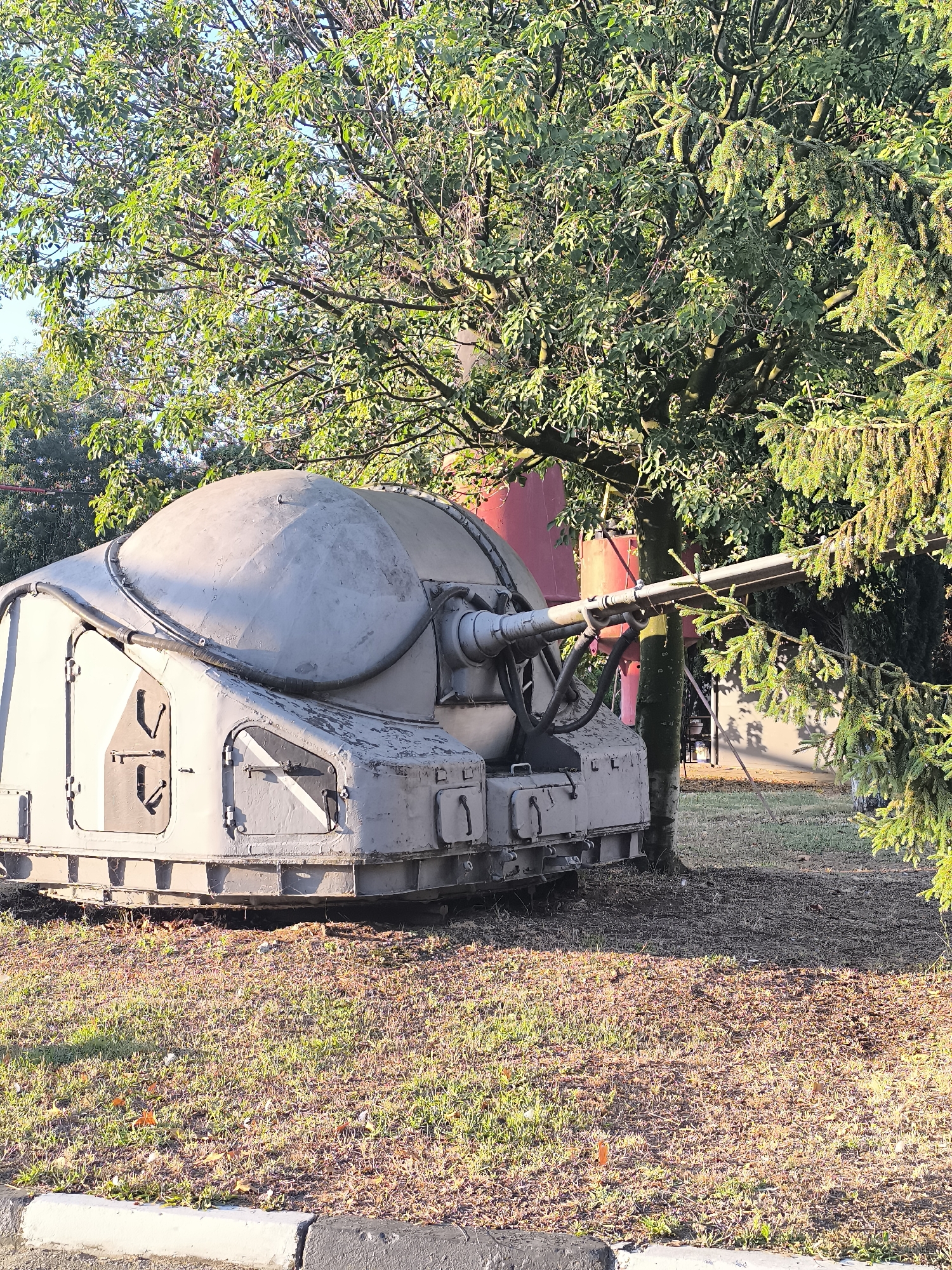 The image shows a static display of an old anti-aircraft gun turret, likely the AK-230 twin-barrel naval gun, situated outdoors in Varna, Bulgaria. The turret is painted a light grey and exhibits signs of wear and tear. It is partially obscured by dense green foliage from trees, with some sunlight filtering through the leaves, casting dappled shadows on the ground. The ground is covered with dry grass and fallen leaves, suggesting it may be autumn. In the background, a tall red cylindrical object is visible behind the turret, along with a portion of a building. The scene appears to be a public space, possibly a park or museum exhibit, given the display of military hardware and the presence of a paved walkway with a curb in the foreground. There are no people visible in the image.