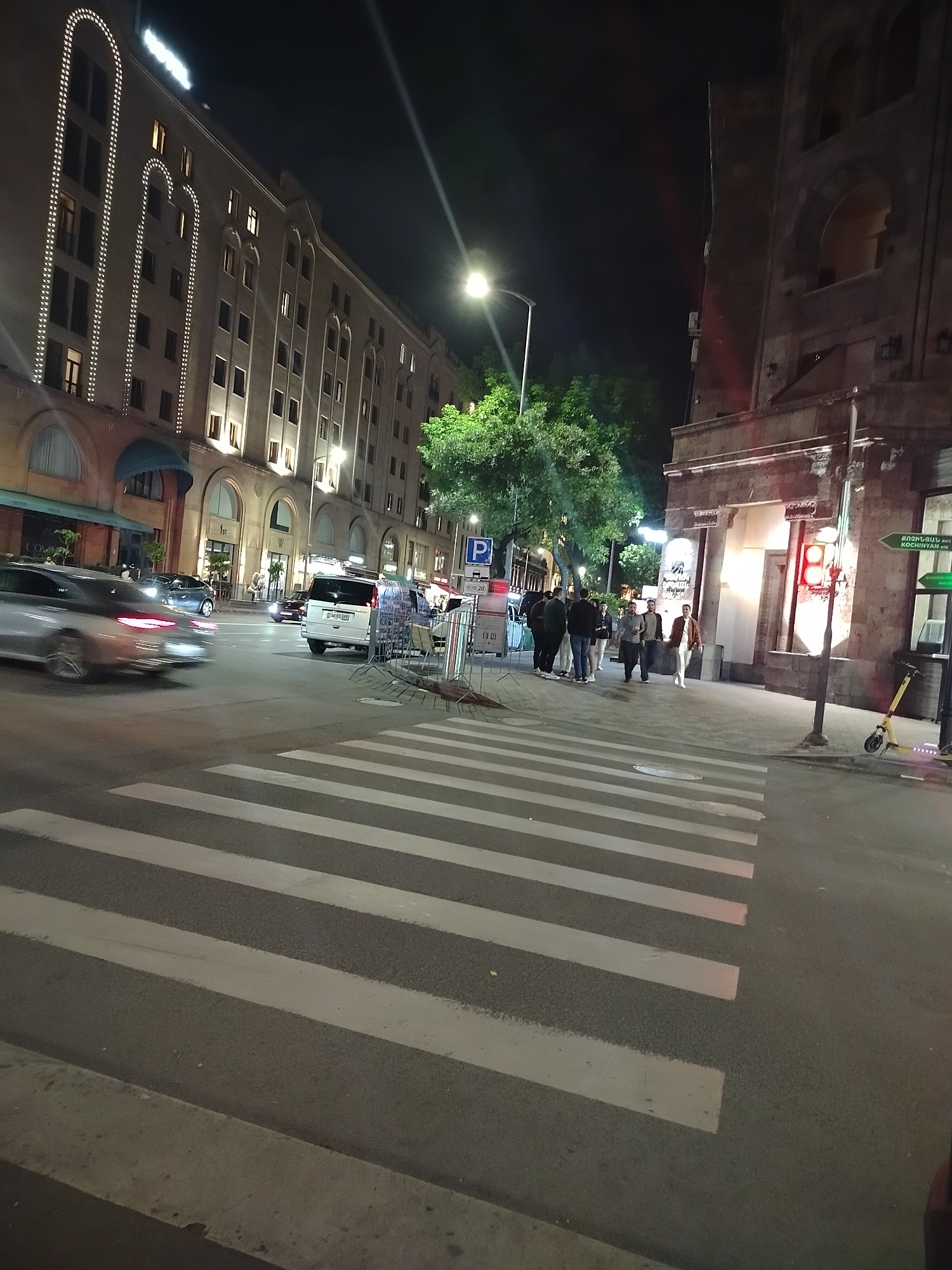 The image depicts a street scene at night in Yerevan, Armenia, specifically at the intersection of Northern Avenue and Abovyan Street, characterized by a crosswalk in the foreground. On the left side of the street, there's a building with a hotel sign on top, adorned with decorative lights. Cars are seen on the road, with one car appearing to be moving at speed, as evidenced by the motion blur. A group of people is gathered on the right side of the crosswalk, seemingly waiting to cross the street. There's a building with a red traffic light and signage on the right side. Streetlights illuminate the scene. The environment appears urban.
