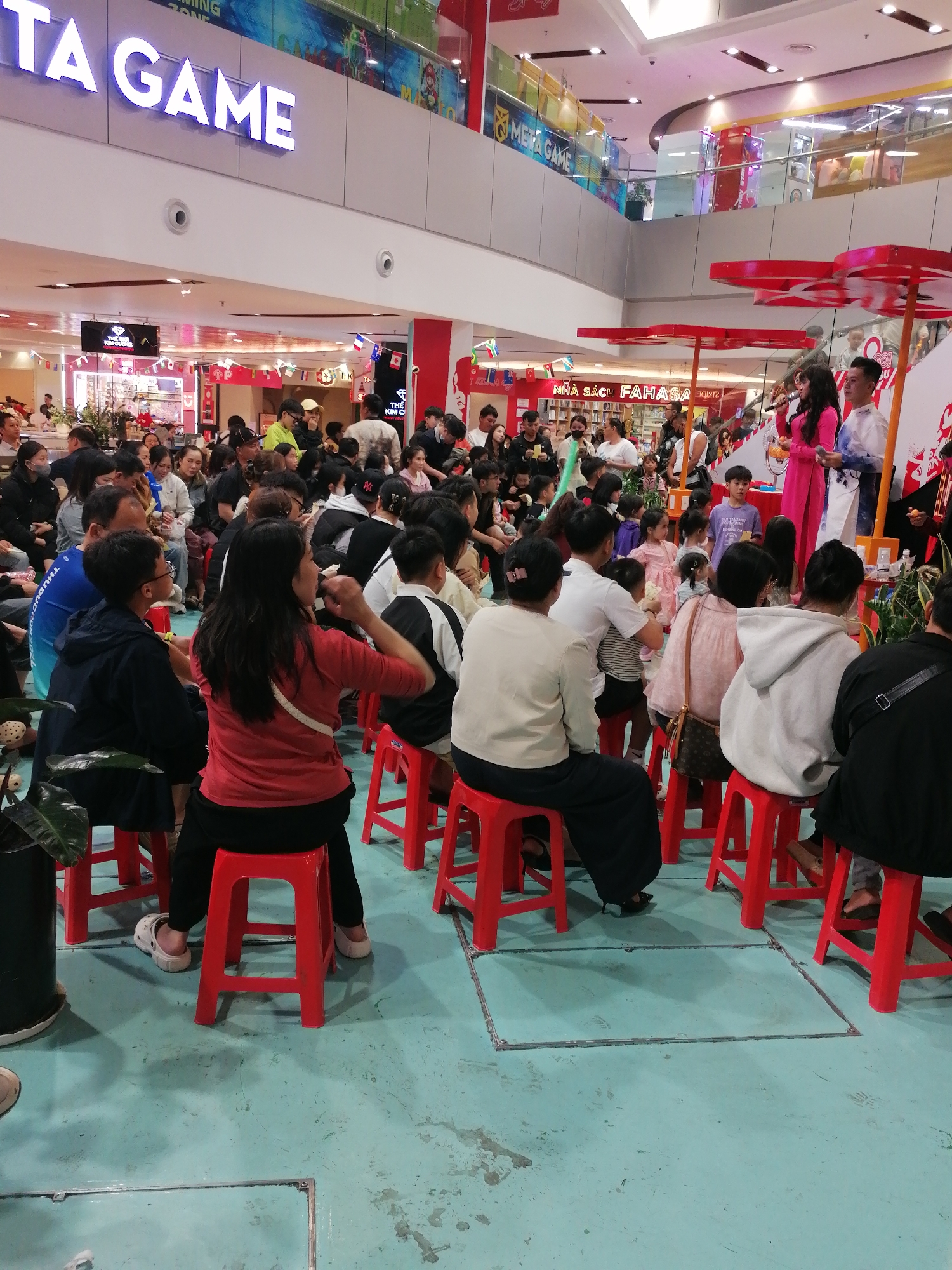 This media file captures a lively indoor event, likely a promotional activity or performance, taking place in a shopping mall. A diverse crowd of people, including adults and children, are seated on bright red stools, facing a central stage area. The scene is brightly lit, suggesting an indoor environment with overhead lighting and possibly natural light filtering from above.
At the front, two performers are visible. A woman in a vibrant pink outfit is holding a microphone, and a man in a white and blue patterned garment stands beside her, appearing to be part of the performance. They are positioned near a display with red decorative elements that resemble large, stylized flowers. Behind them, a section of a bookstore with the visible text "NHÀ SÁCH FAHASA" suggests the event is happening within or in front of a bookshop.
Further in the background, above the crowd, large signs are visible. One sign prominently displays "TA GAME" in large, illuminated white letters, indicating the presence of a game store. Another sign reads "META GAME," further reinforcing the gaming theme of the location. Flags are also visible, adding a festive touch to the setting. The overall atmosphere appears to be engaging and celebratory, with the audience attentively watching the performance. The floor is a light blue, and a plant in a black pot is visible on the left side of the frame. The presence of multiple levels and escalators in the background indicates a multi-story mall.