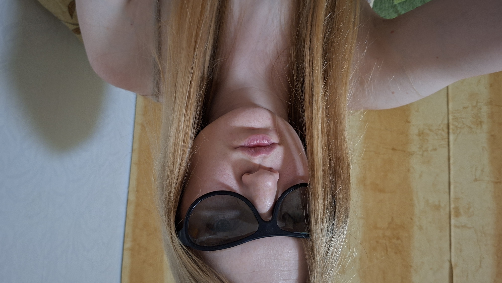 A woman with long, blonde hair and sunglasses is positioned upside down. Her lips are slightly parted. The background consists of a light wall with a patterned surface on the left, and a yellowish-brown wooden or textured surface on the right. There are no visible indications of the location being Большой Букор, Russia.