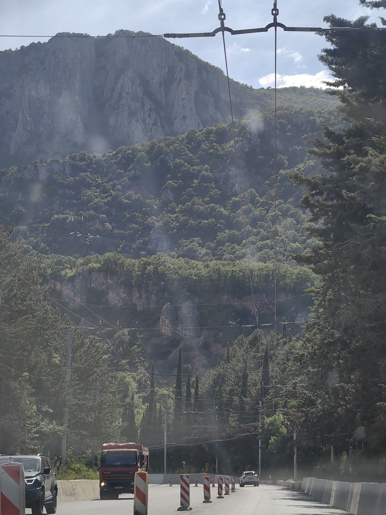 The image depicts a sunlit scene with a road winding through a mountainous, tree-covered landscape. In the foreground, a red truck with a "DAF" logo is visible on the road, its license plate reading "B 678 KA 90". Next to it, a dark SUV is also on the road, partially obscured by construction barriers. The road itself appears to be undergoing some form of construction or repair, indicated by the presence of white and red striped barriers and concrete barriers lining the side.
Behind the road, a dense forest covers the lower slopes of a massive, rugged mountain. The mountain's rocky face is prominent, with visible crevices and sharp angles. Overhead, power lines and what appear to be trolleybus wires crisscross the sky, suggesting an area with public transportation infrastructure. Tall, dark green coniferous trees line the right side of the road, partially obscuring the view of the mountain and sky.
The time of day appears to be daytime, with bright sunlight casting shadows and creating lens flares in the image. The weather seems clear, with some scattered clouds visible in the bright blue sky. The overall impression is of a scenic route through a natural landscape, possibly near a mountainous region or a significant elevation change, with ongoing infrastructure work impacting the immediate road area. No people are clearly visible. The location is identified as Kiparisnoe, Ukraine.