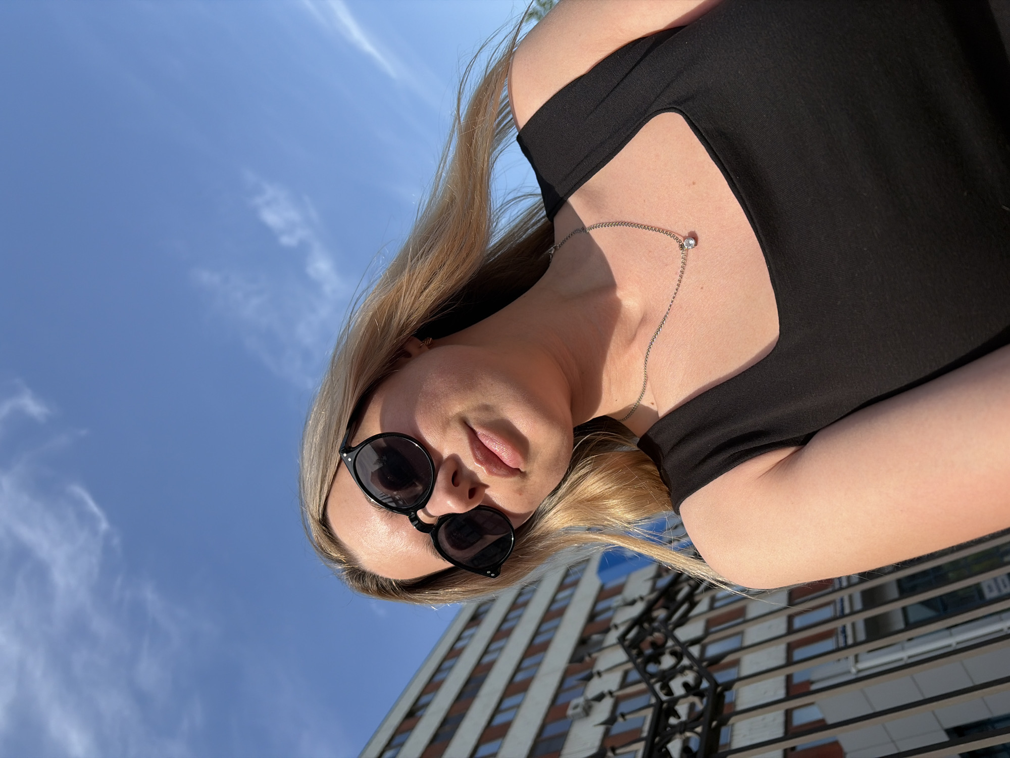 A low-angle, slightly rotated medium shot captures a blonde woman wearing sunglasses and a black top. She is looking directly at the camera with a slightly parted mouth. A delicate silver necklace adorns her neck. In the background is a clear blue sky with wispy clouds. A building with a grid of windows and a dark, wrought-iron fence are partially visible. The scene appears to be in an urban setting, possibly in Chaikovsky, Russia.