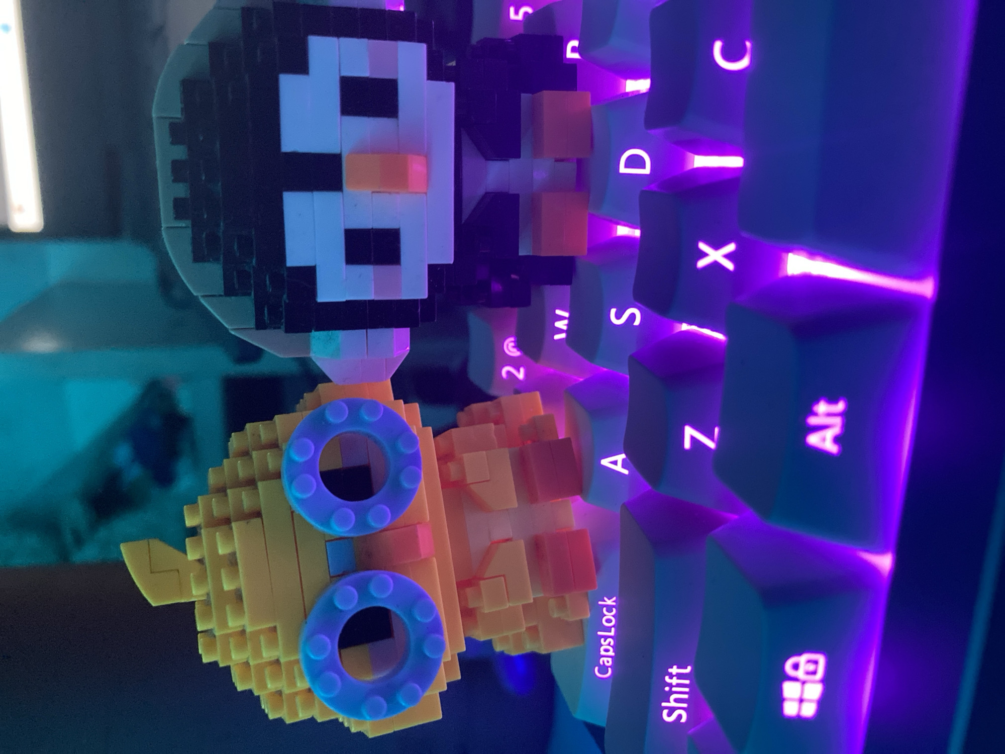 The image shows two pixel art brick figures, a penguin and a yellow object with blue circles, sitting atop a keyboard with purple backlighting. The keyboard keys visible include "C," "D," "X," "S," "Z," "Alt," "Shift," and "Caps Lock." The background is blurry, with a hint of a screen in the upper left. There is no indication of Davao City, Philippines or people in the image.