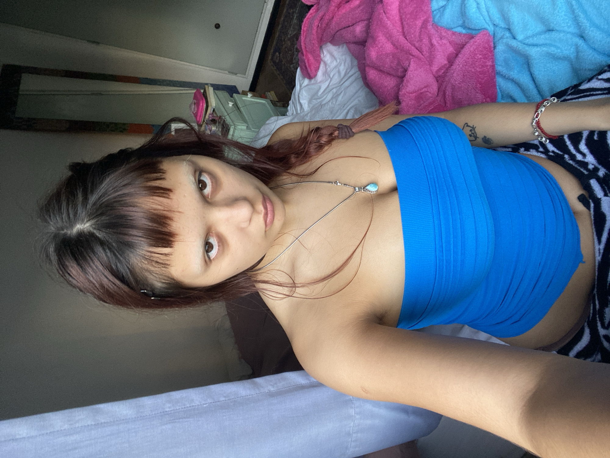 A young woman lies on a bed. She has dark brown hair with bangs and a long braid. She wears a blue bandeau top and black and white patterned pajama pants. She is adorned with a silver necklace with a turquoise pendant. On her arm, there is a tattoo and a red and silver bracelet. The bed is covered with a white sheet, and there are pink and blue towels. In the background, there is furniture, and a mirror on the wall is reflecting the room. It is not possible to determine the location of Buenos Aires, Argentina from the image.