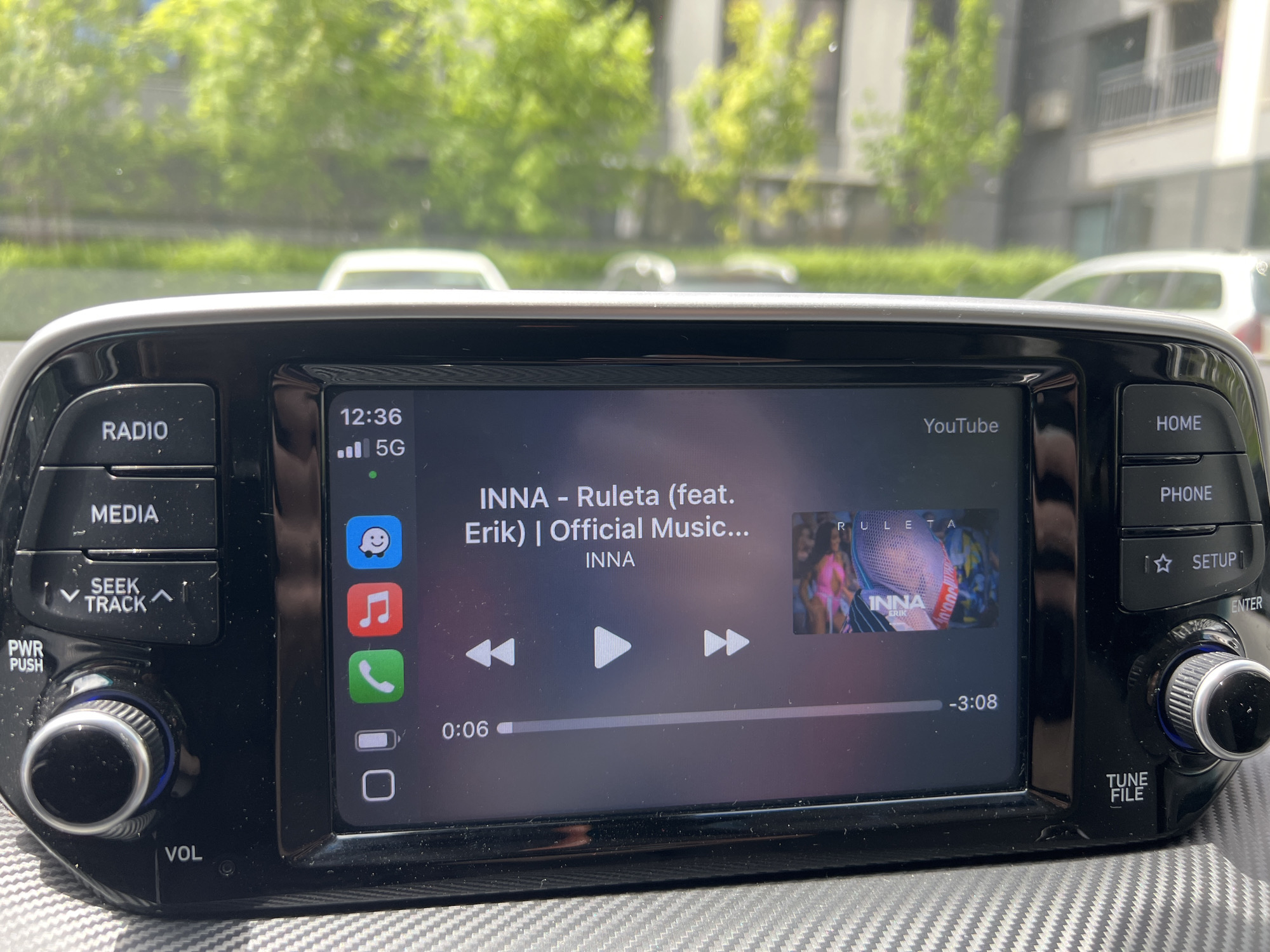 The image shows the infotainment screen of a car, displaying a YouTube video of the song "Ruleta" by INNA featuring Erik. The screen also indicates that the car is connected to 5G network. The time is 12:36. The display has icons for messages, music and a phone. The background outside the car window shows trees and buildings, suggesting an urban environment. There is no way to confirm that this picture was taken in Burgas, Bulgaria from the image.