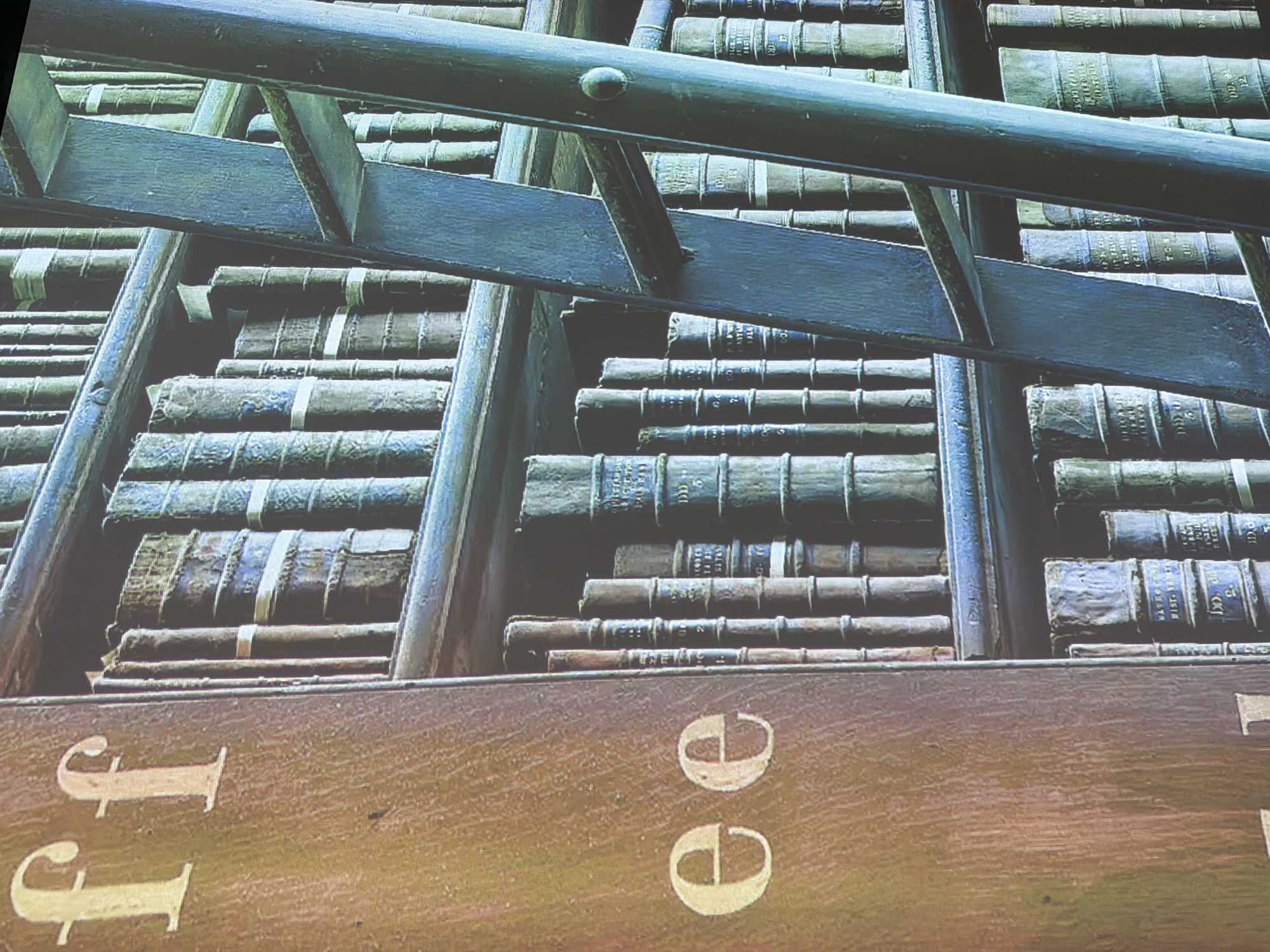 The image displays a close-up of a bookshelf filled with old, leather-bound books. A wooden ladder, painted a muted blue, leans against the shelves, suggesting a library or study. The books are tightly packed, and some have visible titles or markings on their spines, though they are mostly illegible due to the resolution and angle. The lighting suggests an indoor environment, possibly dimly lit, with a warm glow on the wooden elements and a cooler tone on the ladder and some of the book bindings. There are no people visible in the frame. The image appears to be a photograph of a physical scene, with the textures of the books, wood, and ladder being prominent. The location is not specified within the image itself and cannot be determined without additional information.