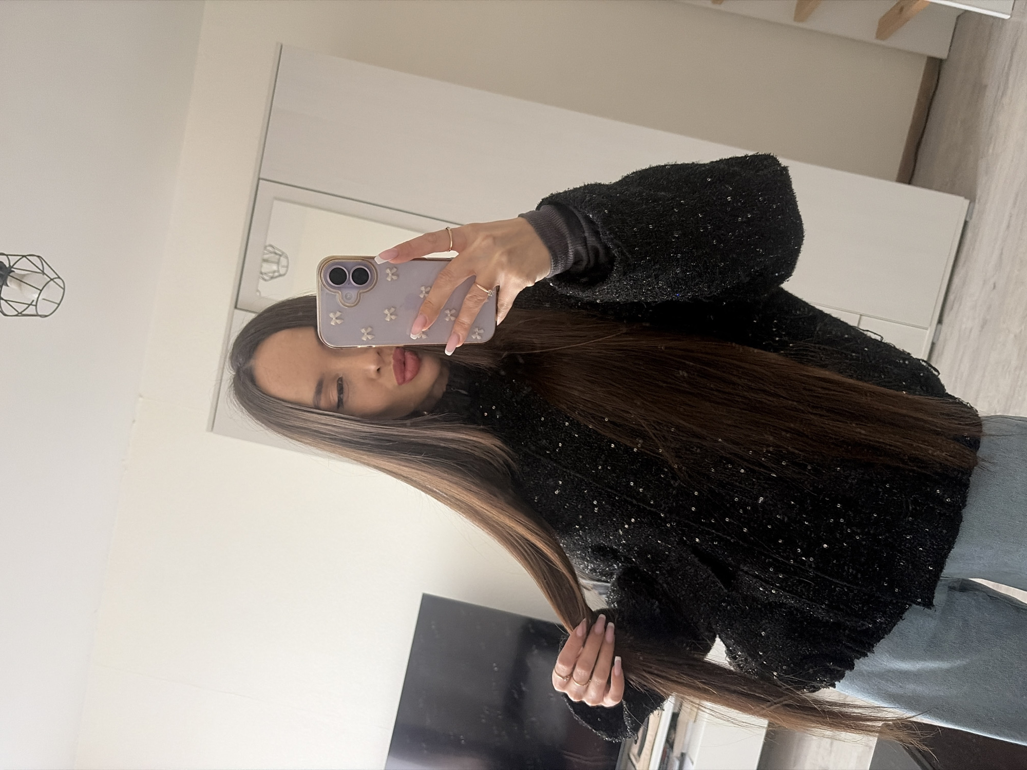 A woman is taking a selfie in an indoor environment. She is holding a light purple smartphone in her right hand. The woman is wearing a black textured sweater over a gray top and light blue jeans. Her long, dark hair falls over her sweater. The background includes white walls, a white wardrobe, a black TV, and a wooden floor. The location is undetermined and cannot be identified as Saint Petersburg, Russia, from the given information.