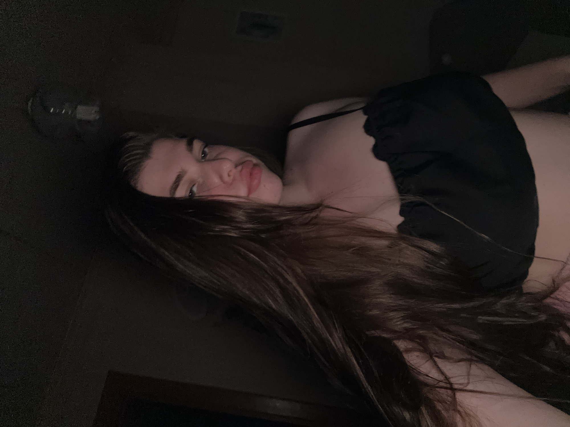 A medium shot depicts a woman with long, brown hair in an indoor setting. She is wearing a black top with straps. The background appears dark and includes a wall and a lighting fixture. The woman is looking at the camera with a slight pout. Location: Unable to determine if the location is in the city of Мещёры, Russia.