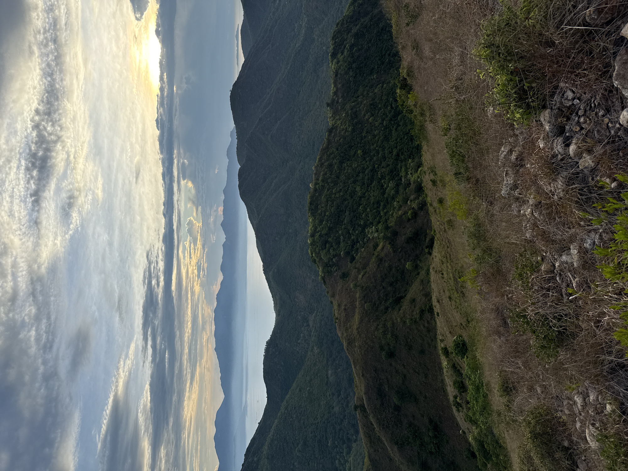 The image shows a landscape in Bac Nha Trang Ward, Vietnam. The foreground presents a steep hillside covered with dry grass, shrubs, and some rocks. Moving into the midground, a series of tree-covered mountains cascade into the distance, varying in shade from dark green to hazy blue. In the background, a body of water is visible between the mountains, and above it, the sky is filled with striated clouds, illuminated by the sun breaking through.