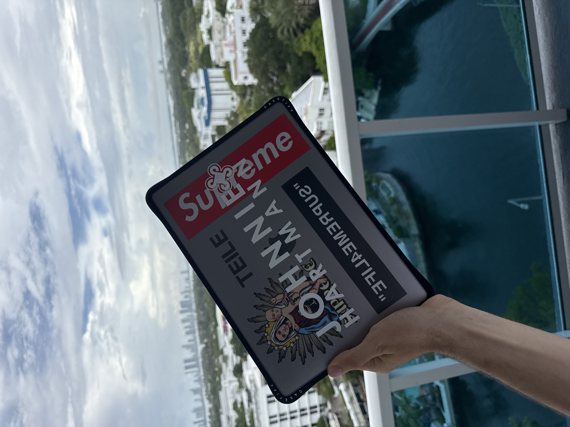 A person is holding a gray case with a "Supreme" logo in front of a high-rise view in Miami Beach, United States. The case features additional text and an image of a woman holding a baby. The background includes white buildings, trees, water, and a bridge. The sky is cloudy. The person is holding the case in their right hand. A window frame is visible on the right side of the image.