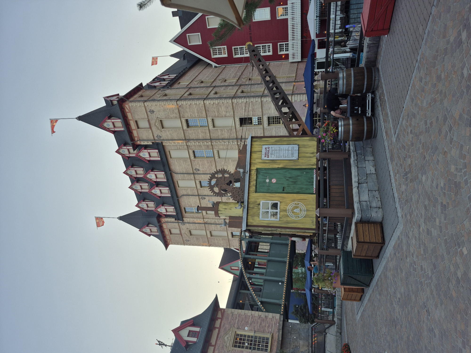 The image depicts an outdoor scene with several buildings in a European-style architectural setting, likely within the Europa-Park in Rust, Germany. The main building is a tall, multi-story structure with a light-colored brick facade, a dark pitched roof with numerous dormer windows, and red trim. To the left, a smaller building with a red roof and brick facade is visible. To the right, a prominent red building with white-framed windows and a white balcony stands out.
In the foreground, a distinctive yellow structure resembling an old industrial building or a prop from a themed attraction is present. It features a large green door, a window, and a decorative crane arm extending from its side. Several wooden crates and barrels are positioned around this structure, along with planters containing flowers. Two blue umbrellas provide shade over a seating area behind a dark railing.
A man wearing a black t-shirt and pants is bending over equipment placed between two wooden barrels, appearing to be setting up or adjusting something. The ground is paved with interlocking stones. Flags are visible atop some of the buildings, suggesting a festive or themed environment. The sky is a pale, hazy blue, indicating either early morning or late afternoon. The overall atmosphere suggests a tourist or entertainment district.
