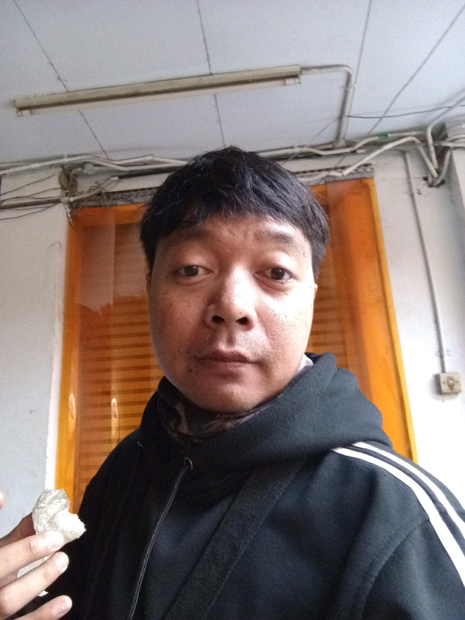 A medium shot selfie shows a person holding a piece of food in their left hand. The person has dark hair and is wearing a black hooded jacket with white stripes on the shoulders. The background consists of a white wall, an orange curtain or blind, and a ceiling with electrical conduits and lighting. The image appears to have been taken indoors, possibly in a workshop or storage area. There is no visual information within the image to suggest the location is Bandung City, Indonesia.