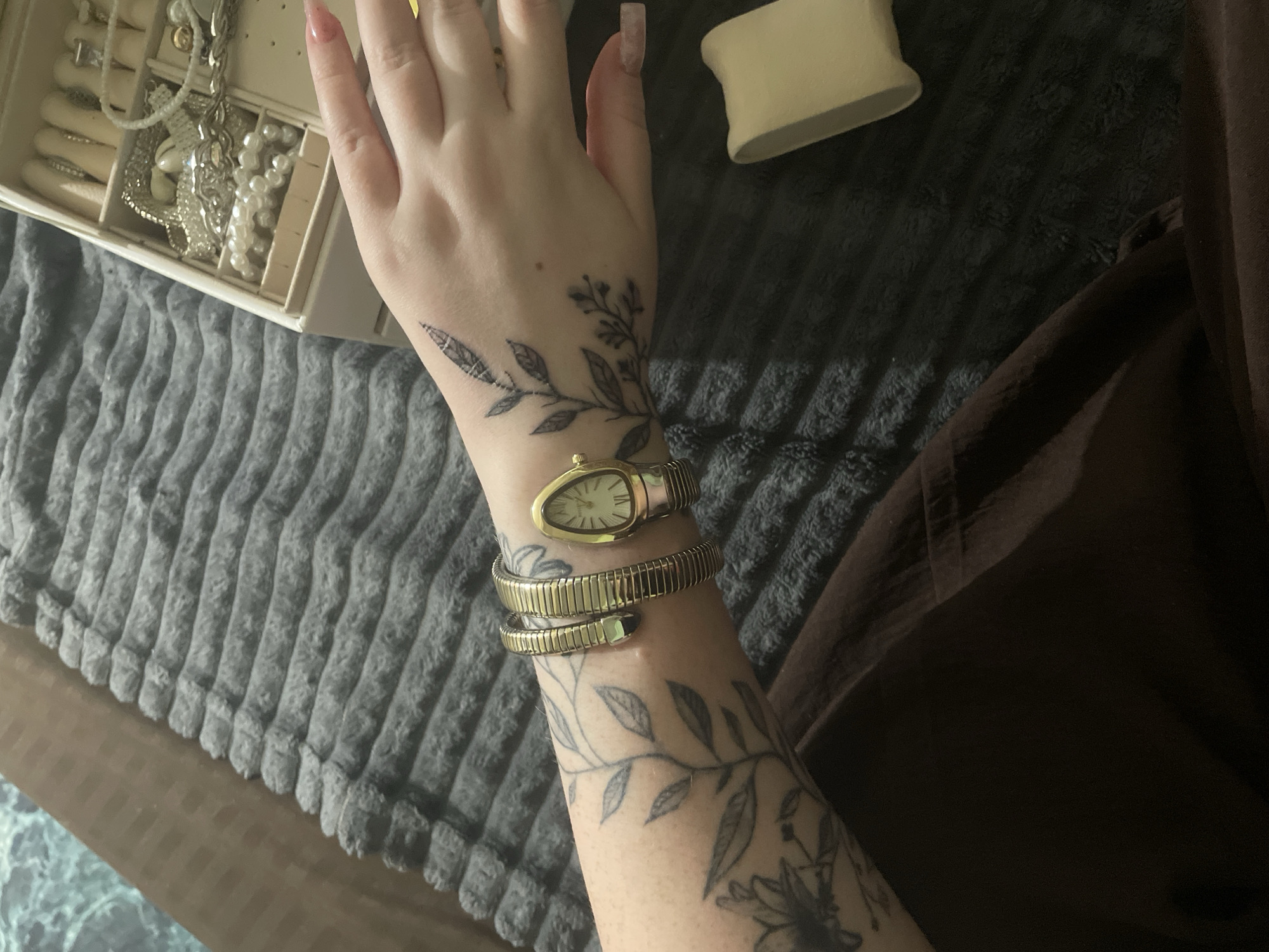 In Линёвка, Russia, a person's left forearm and hand are visible, resting on a dark grey, textured blanket. The forearm is adorned with floral and leafy tattoos. On the wrist, a gold, snake-shaped bracelet is worn, and next to it, a gold watch with a white oval face. The hand, positioned palm-down, also has a delicate leaf tattoo extending from the wrist onto the back of the hand. A jewelry box, filled with various silver necklaces, bracelets, and pearls, is partially visible in the upper right corner. The background consists of the grey blanket and a portion of a dark brown garment.