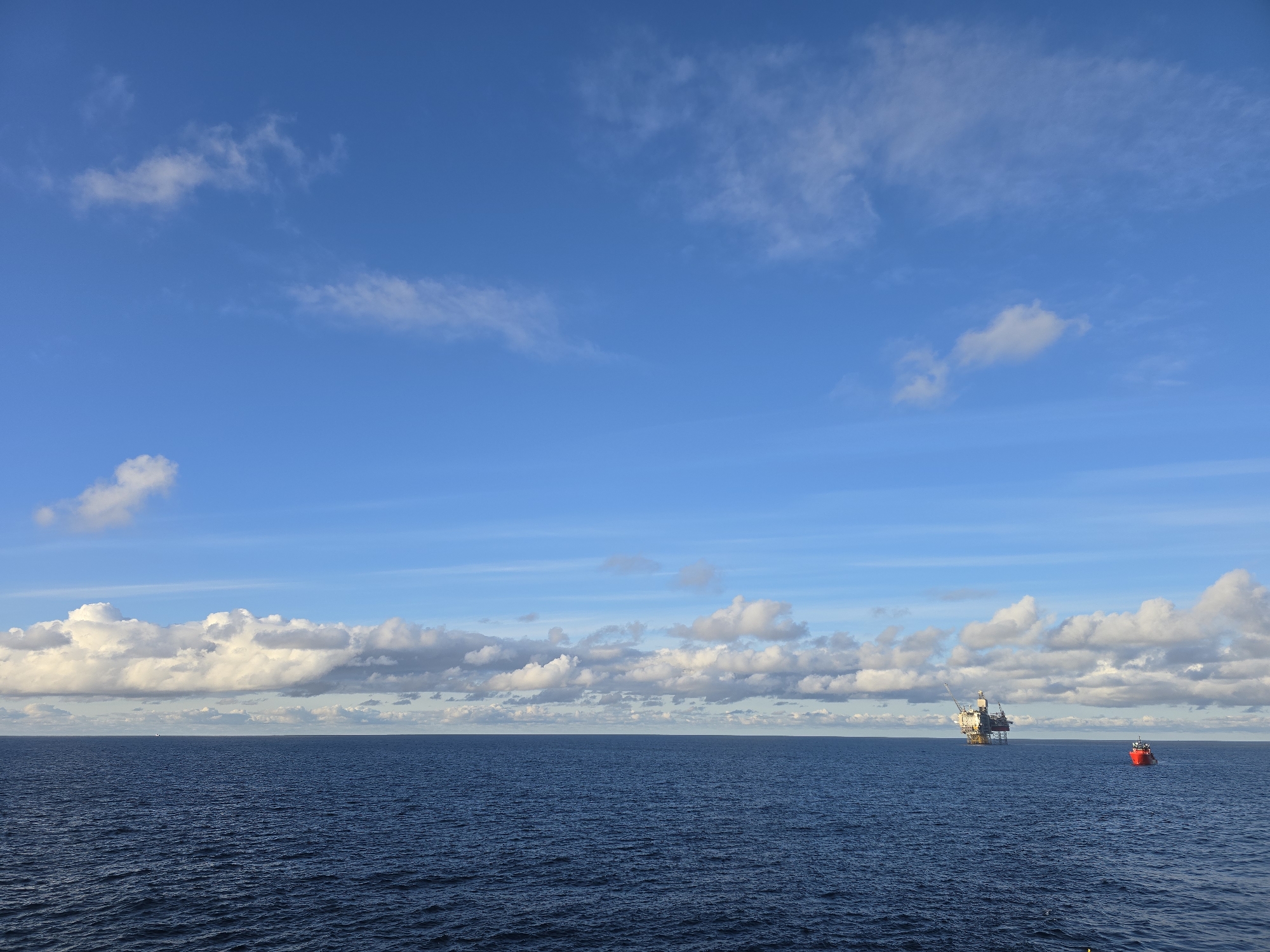 The image depicts a seascape under a blue sky with scattered clouds. A body of dark blue water extends to the horizon. In the distance, a large offshore oil platform is visible. To the right of the platform, a red boat is present on the water. The location is the city of Unknown, Unknown.