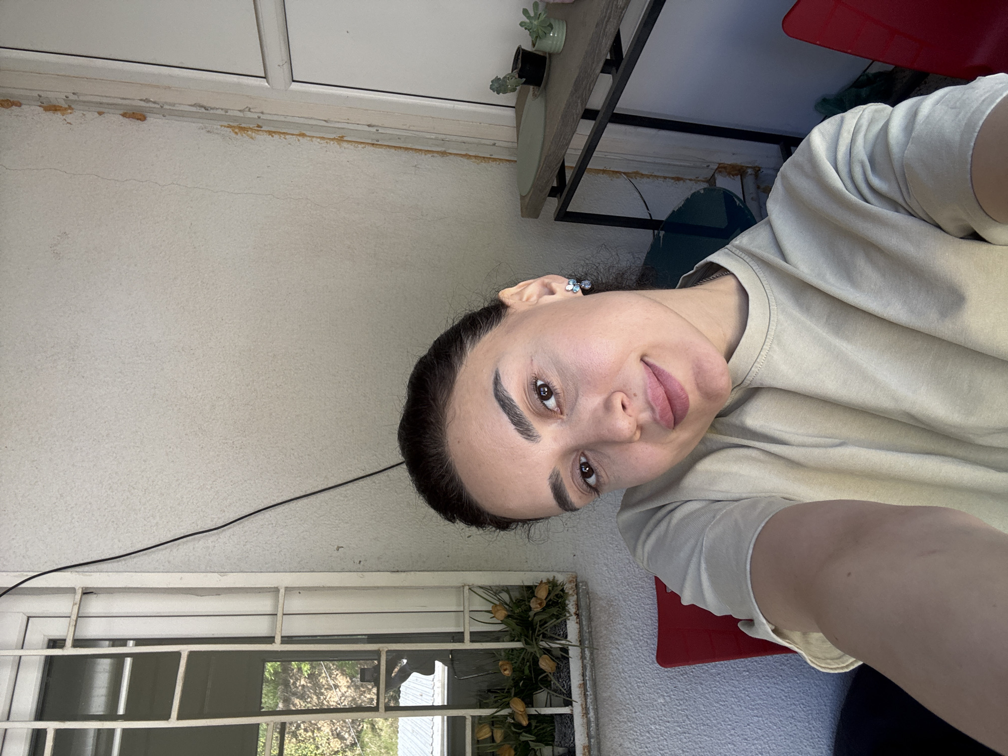 A person is pictured in what appears to be an outdoor setting in Tbilisi, Georgia. The subject, a woman with dark hair and distinct eyebrows, is wearing a light-colored T-shirt and earrings. She is positioned at an angle to the camera, with her face partially visible. Behind her, there is a white wall with a window frame and bars, through which some greenery is visible. A shelf with small potted plants is located above her. The scene is partially lit by natural light. A red surface, possibly a chair or pad, is visible.
