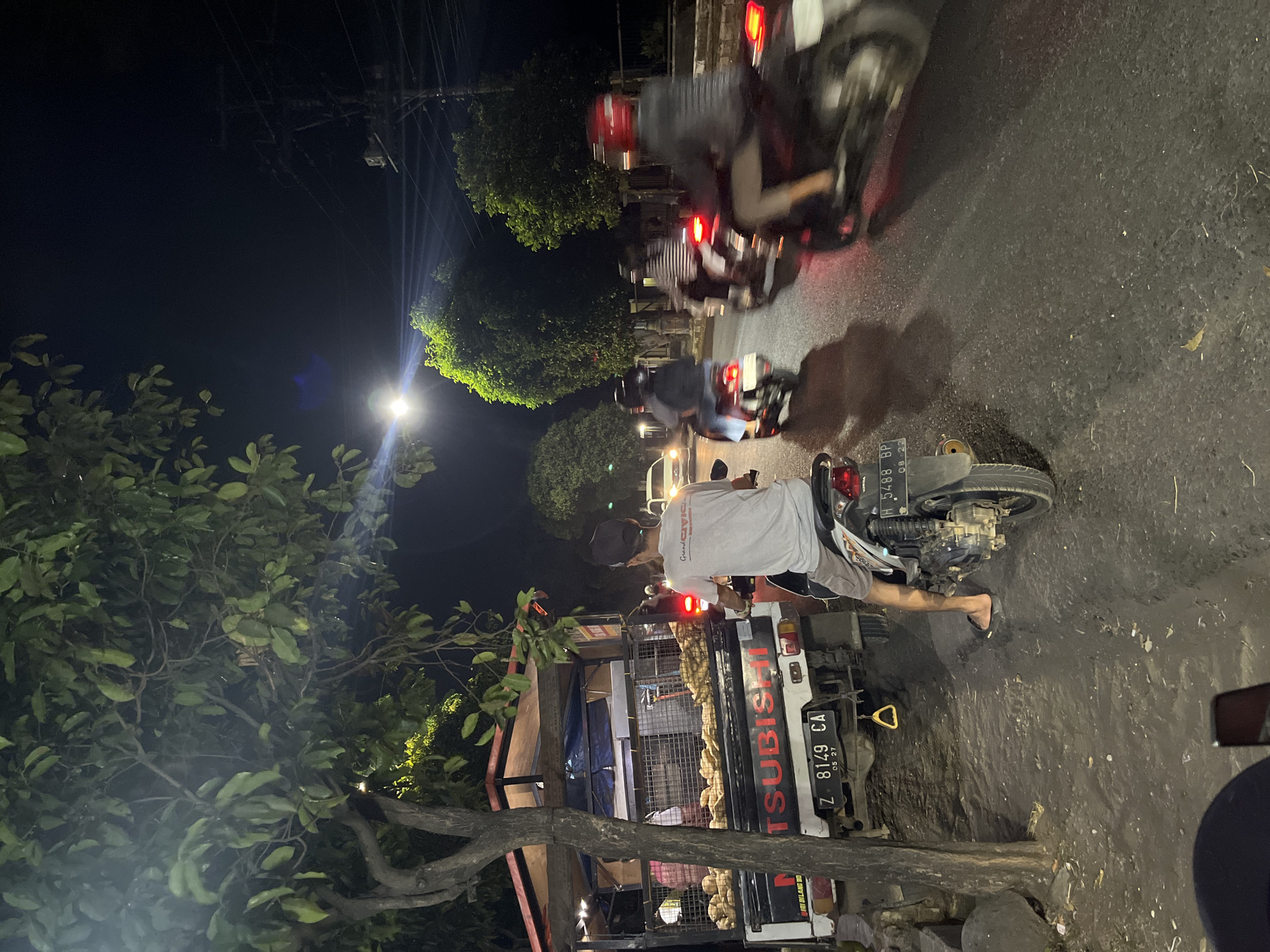 This is a nighttime scene on a street in Pecangaan, Indonesia. A white Mitsubishi truck, laden with produce, is parked on the side of the road. A man wearing a gray t-shirt and shorts is sitting on a scooter behind the truck, facing away from the camera. The scooter has a license plate that reads "H 5488 BP". Several other people on scooters are visible on the road, some of whom are moving and others who appear to be stationary. The street is paved and appears to be slightly wet. Trees line the side of the road, and a street light illuminates the scene. The overall impression is one of a busy street during evening hours in a Indonesian town.
