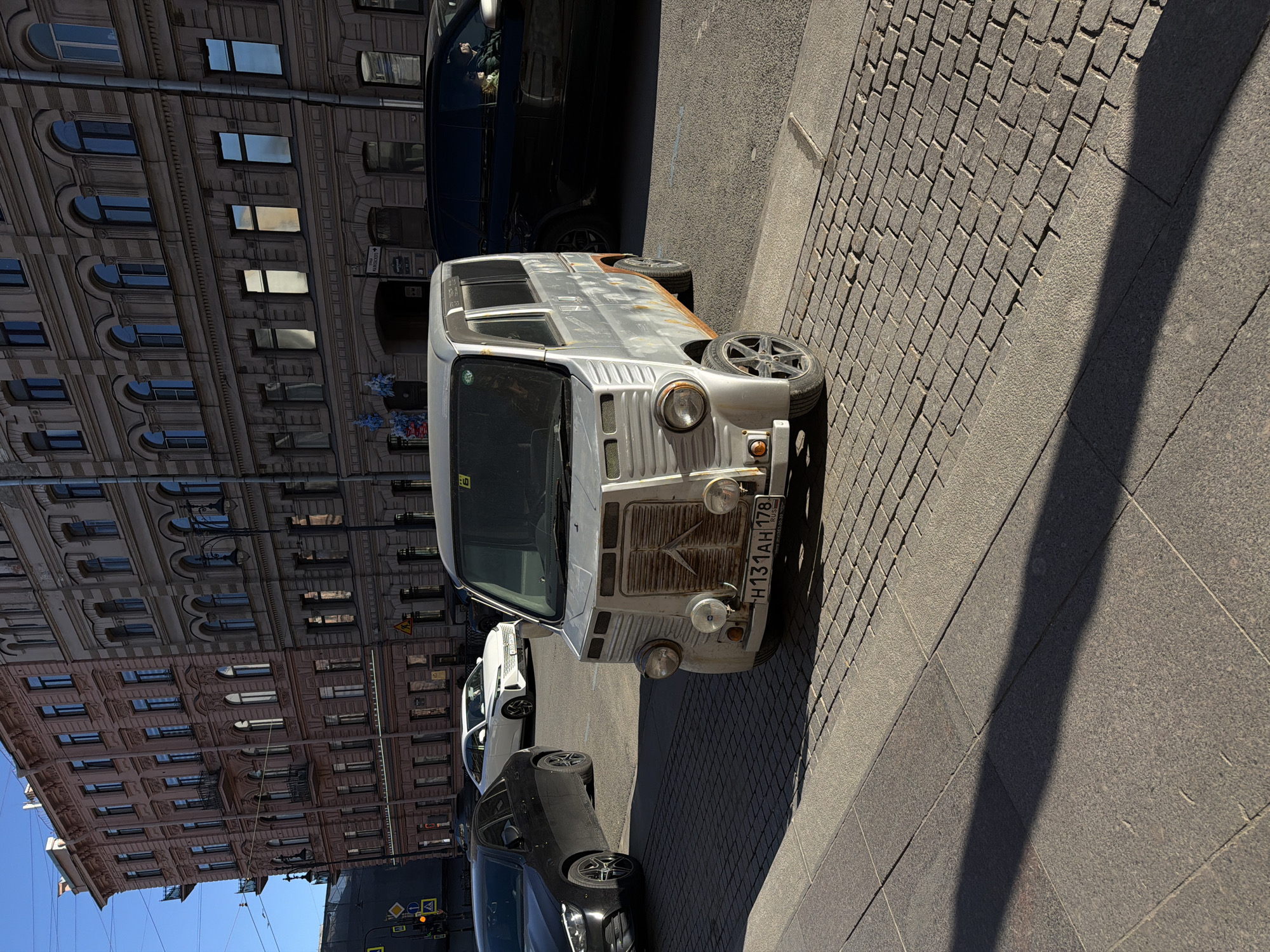 The image captures a street scene in Saint Petersburg, Russia. The primary subject is a light-colored, vintage van with visible signs of wear and rust, parked alongside a cobblestone street. The vehicle's license plate, "H131 AH 178," is partially visible. Several other vehicles, including a dark-colored sedan and a white car, are parked nearby. A multi-story building with ornate architectural details and numerous windows forms the backdrop, partially visible against a clear, blue sky. The scene is well-lit, suggesting a sunny day.