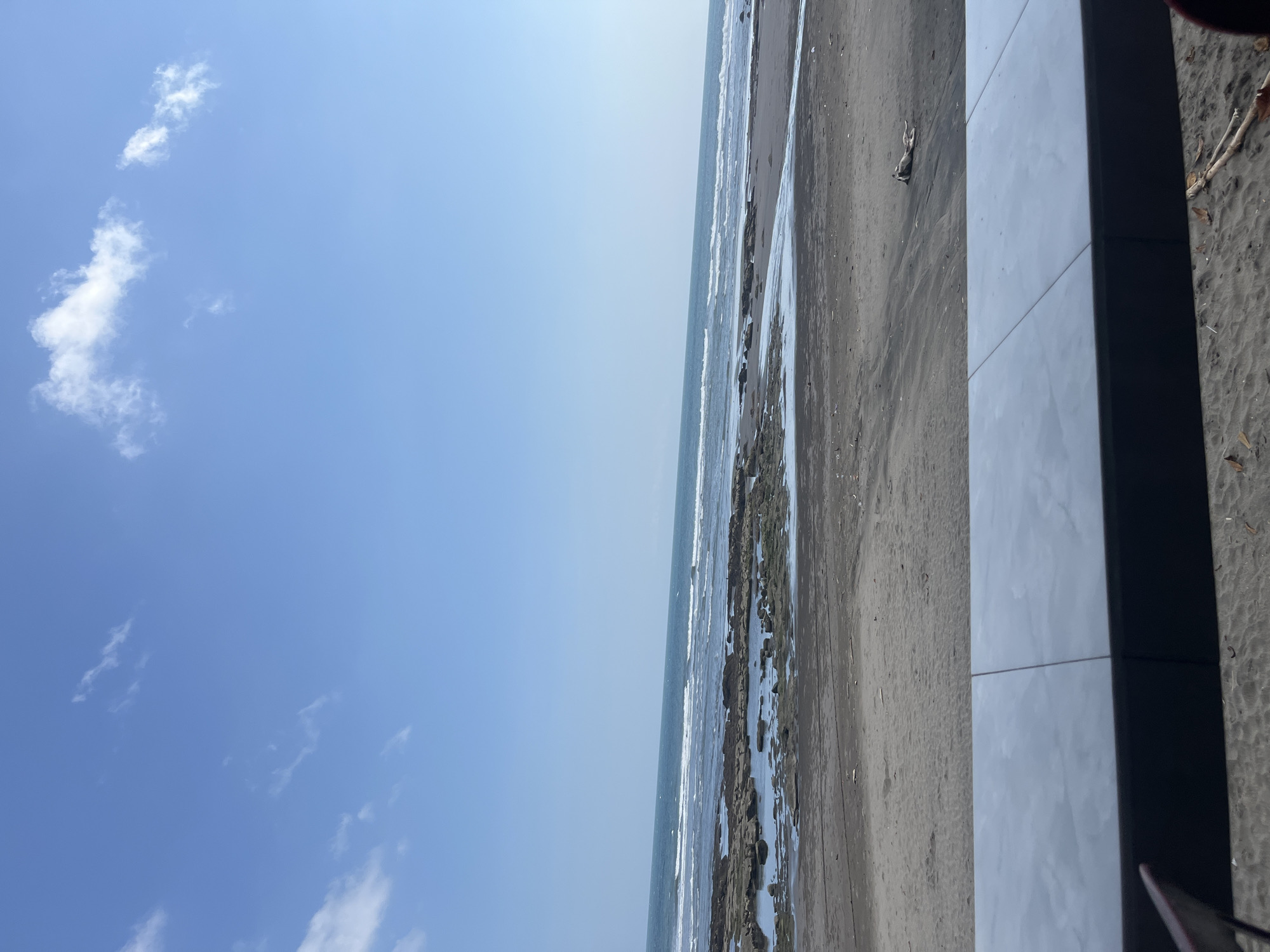 This is a photograph of a beach in Pekutatan, Indonesia. The sky is a clear blue with a few scattered white clouds. The ocean is a deep blue, with gentle waves breaking on the shore. The beach is covered in dark sand, with a scattering of rocks and driftwood. In the foreground, there is a tiled ledge, suggesting this is a vantage point overlooking the beach, perhaps a cafe or seating area. The tide is out, revealing shallow pools of water among the rocks. The overall impression is one of a serene and natural coastal landscape.