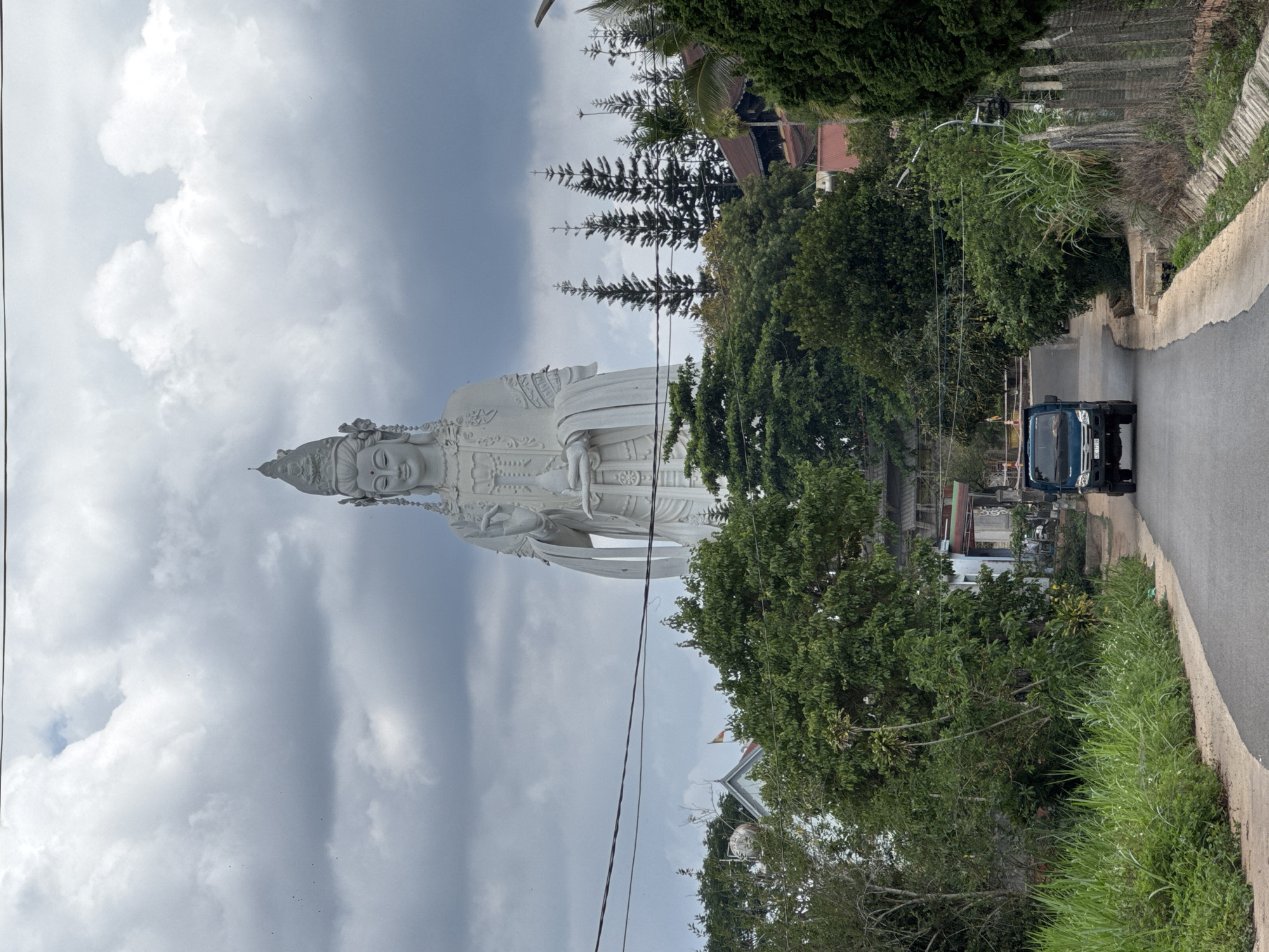 The image shows the large white statue of Guanyin in the city of Nam Ban, Vietnam, positioned against a cloudy sky. The statue is located next to a road with a blue truck on it and surrounded by trees. There are power lines in the foreground. The foliage appears dense, and there is some evidence of nearby buildings visible through the trees.