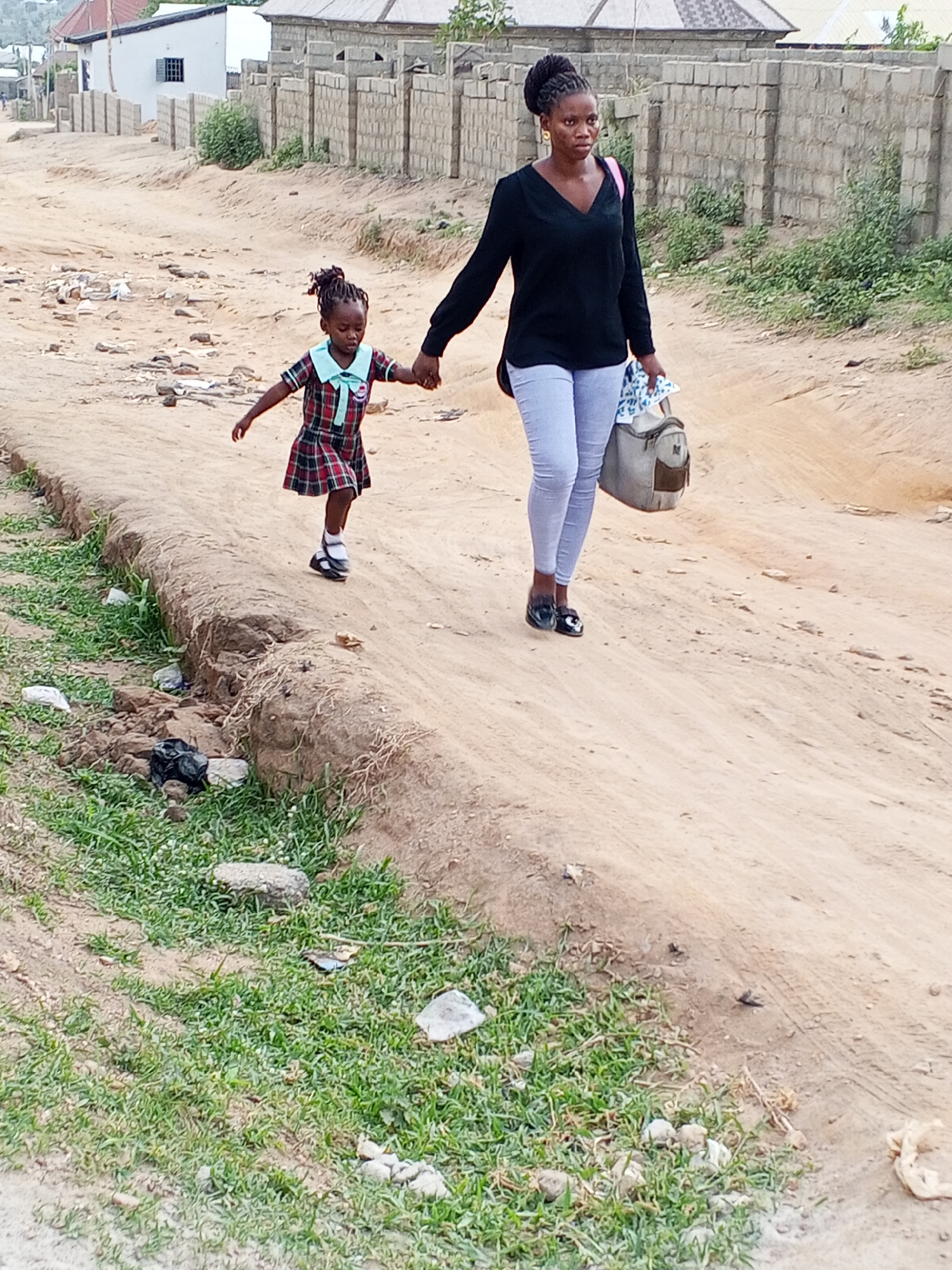 A mother and daughter are walking hand-in-hand on a dirt road in an urban area. The mother is wearing a black long-sleeved shirt, light blue jeans, and black shoes. She has her hair up in a bun. A pink strap of a bag is visible on her shoulder, and she is carrying a gray bag in her other hand. The daughter is wearing a red and green plaid dress with a light blue collar and black shoes. Her hair is styled in small braids with colorful beads. The road is unpaved and has patches of grass along the side. There is trash scattered around. In the background, there are houses and a brick wall. The location appears to be in a residential neighborhood of Unknown, Unknown.
