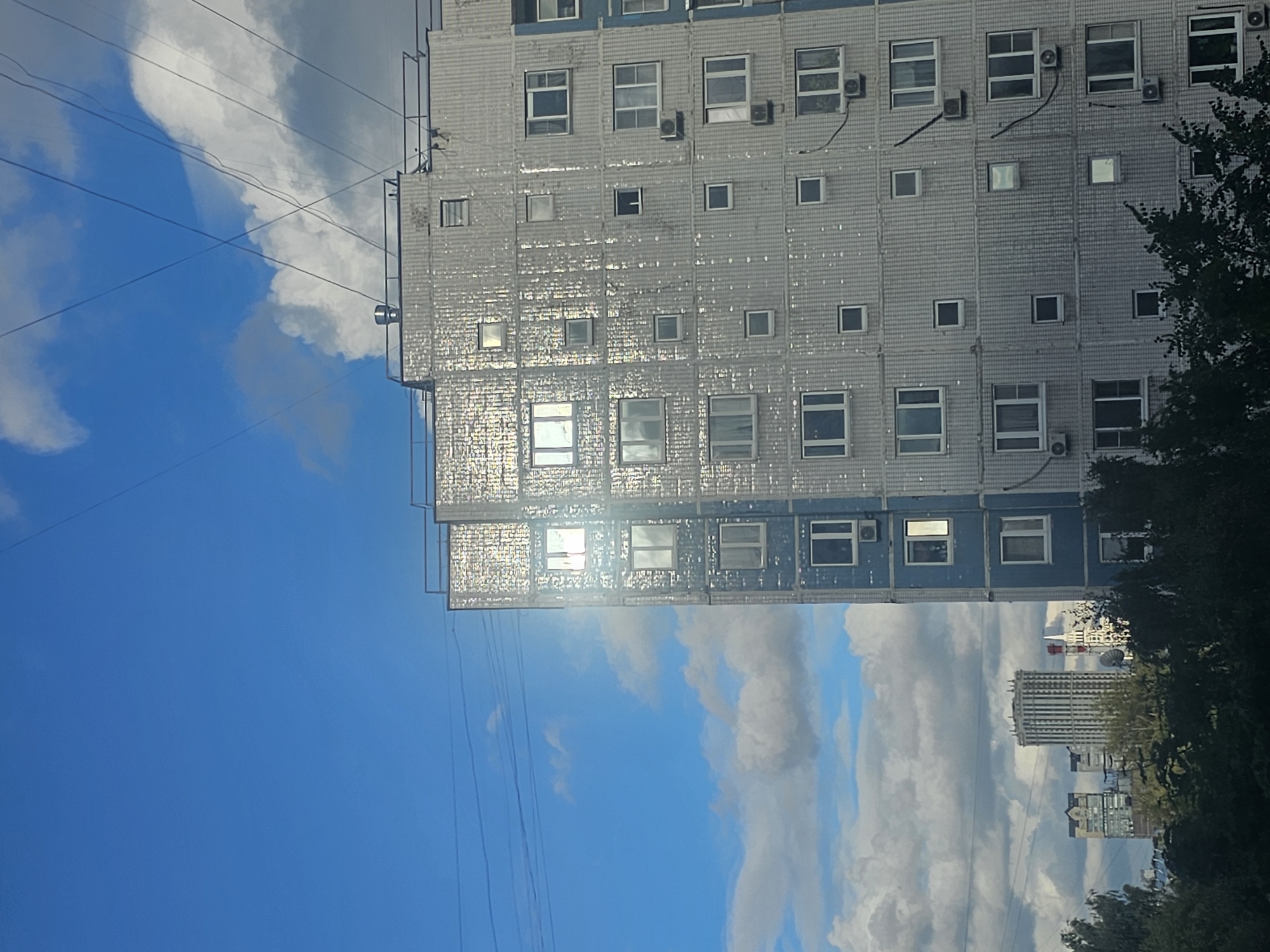 An eye-level, full shot captures a residential building in Khimki, Russia, under a bright, cloudy sky. The building, primarily white with blue accents, dominates the right side of the frame. Its facade is covered in a textured, reflective material, causing a strong glare from the sun. Numerous windows, some with air conditioning units below them, are visible across multiple floors. The building's top features a railing and a ventilation pipe. In the distance, other buildings, including a prominent multi-story structure with a distinct architectural style, are partially obscured by trees. Overhead, power lines crisscross the sky. The sky itself is a vibrant blue, with large, puffy white clouds interspersed with darker, shadowed clouds. The overall impression is of an urban landscape on a sunny, yet somewhat overcast day.