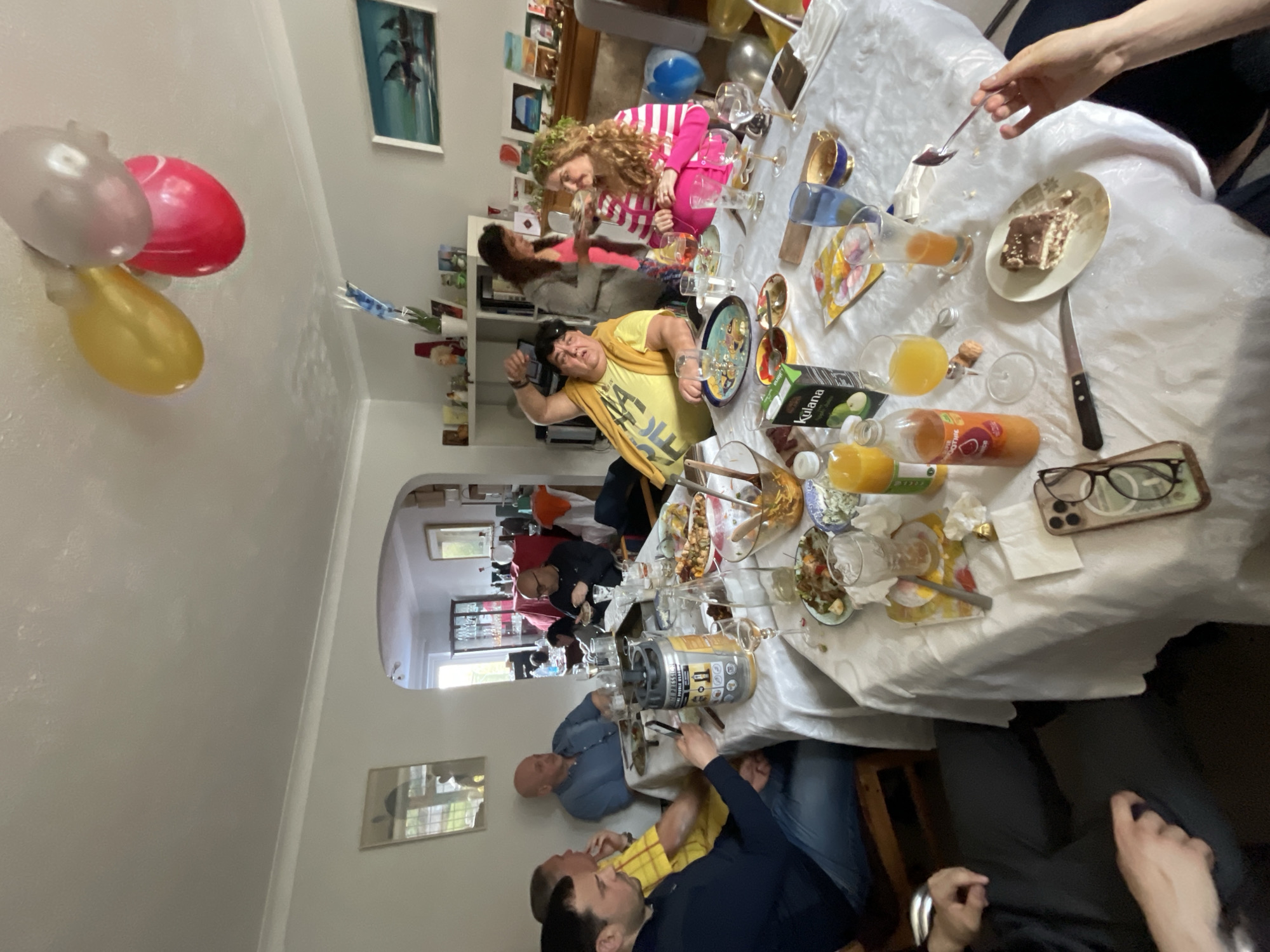 A slightly rotated, high-angle shot captures a lively gathering indoors, likely a celebratory event. The setting is brightly lit and appears to be within a residential home. At the center is a long table covered with a white tablecloth. The table is laden with food, drinks, bottles of juice, used plates, glasses, and cutlery, suggesting a recent meal or ongoing festivity. A piece of cake sits on a plate, next to a knife and a pair of eyeglasses.
Multiple individuals are present. Some are seated around the table, engaged in conversations and interactions, while others are standing or moving around. One person in a yellow shirt stands and raises their fist. There are balloons visible in the room, adding to the celebratory atmosphere.
Through an arched doorway, another room can be seen, filled with more people and suggesting the home is quite large. Paintings adorn the walls. Given the available information, determining the exact location in Maidstone, United Kingdom is impossible.