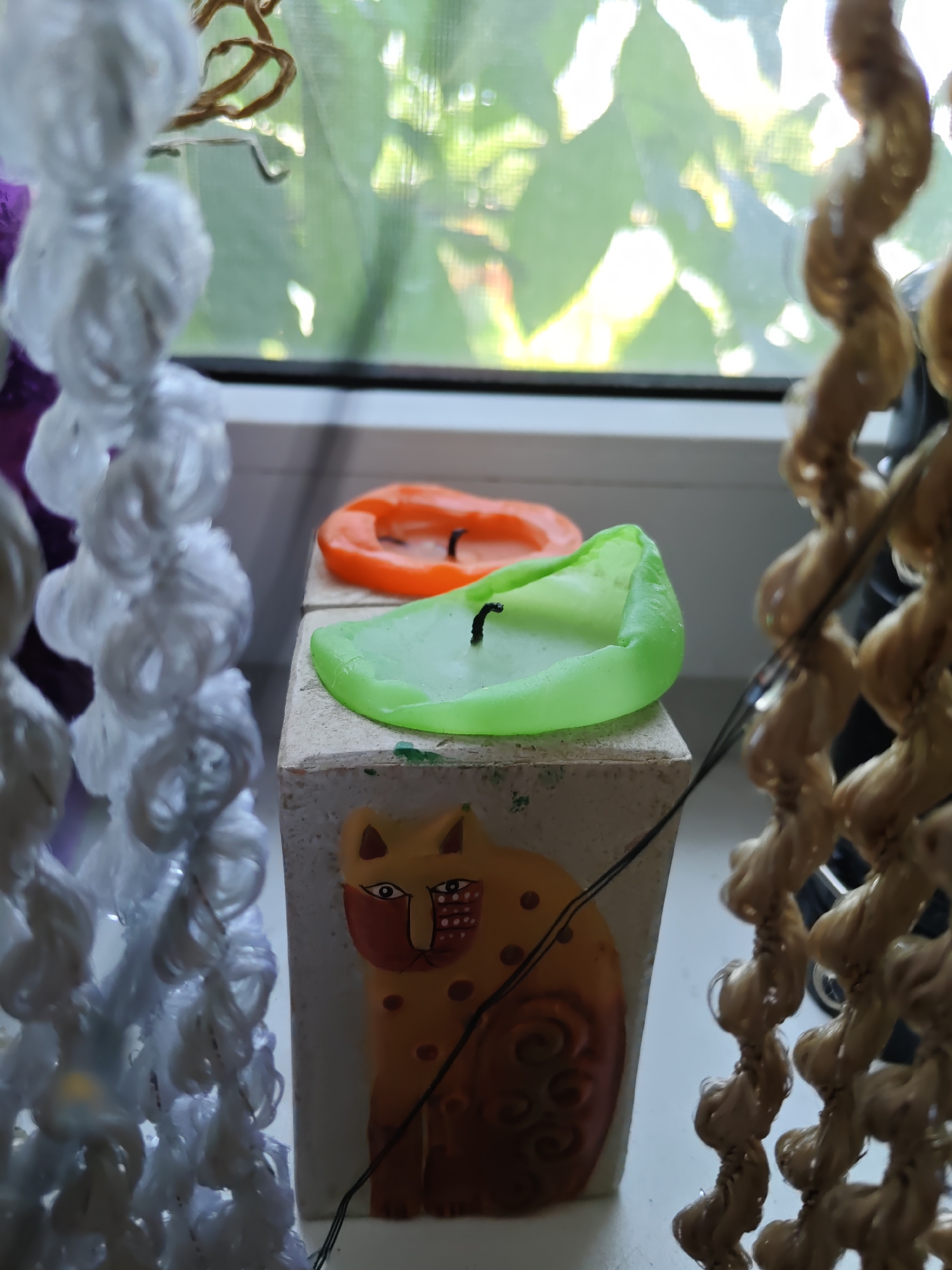 In Rostov-on-Don, Russia, on a windowsill, two melted candles sit atop a wooden block with a painted cat design. One candle is orange, and the other is green, both showing signs of having been lit. The background reveals a window with a blurred view of foliage outside. To the left and right of the scene, decorative string elements with a twisted texture hang, obscuring parts of the view. The lighting suggests it is daytime.