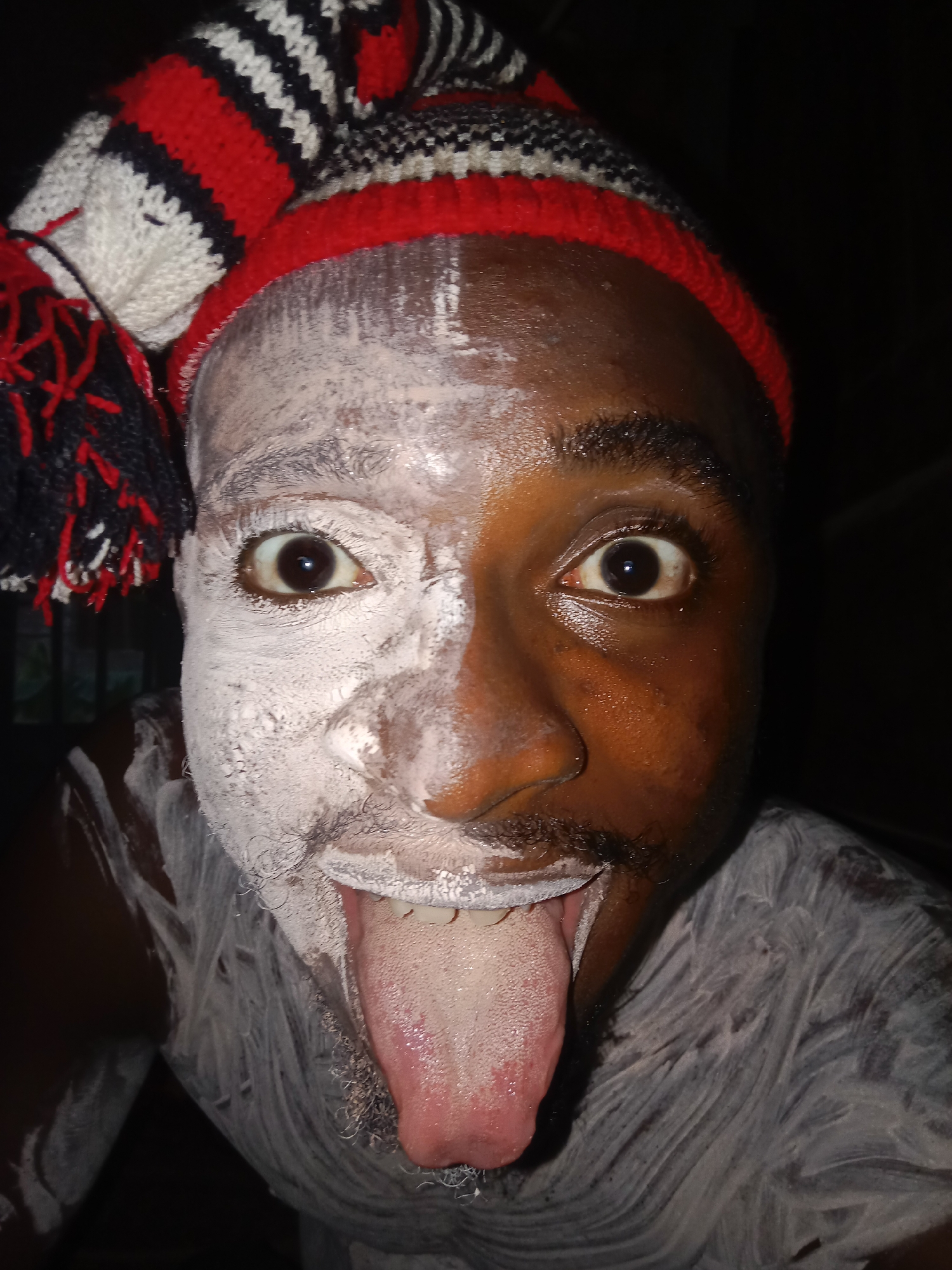 A close-up shot shows a man with half of his face covered in white paint or powder. He is wearing a red, white, and black striped hat with tassels. His tongue is sticking out, and his eyes are wide open. The background is dark and out of focus. The location is Unknown, Unknown.