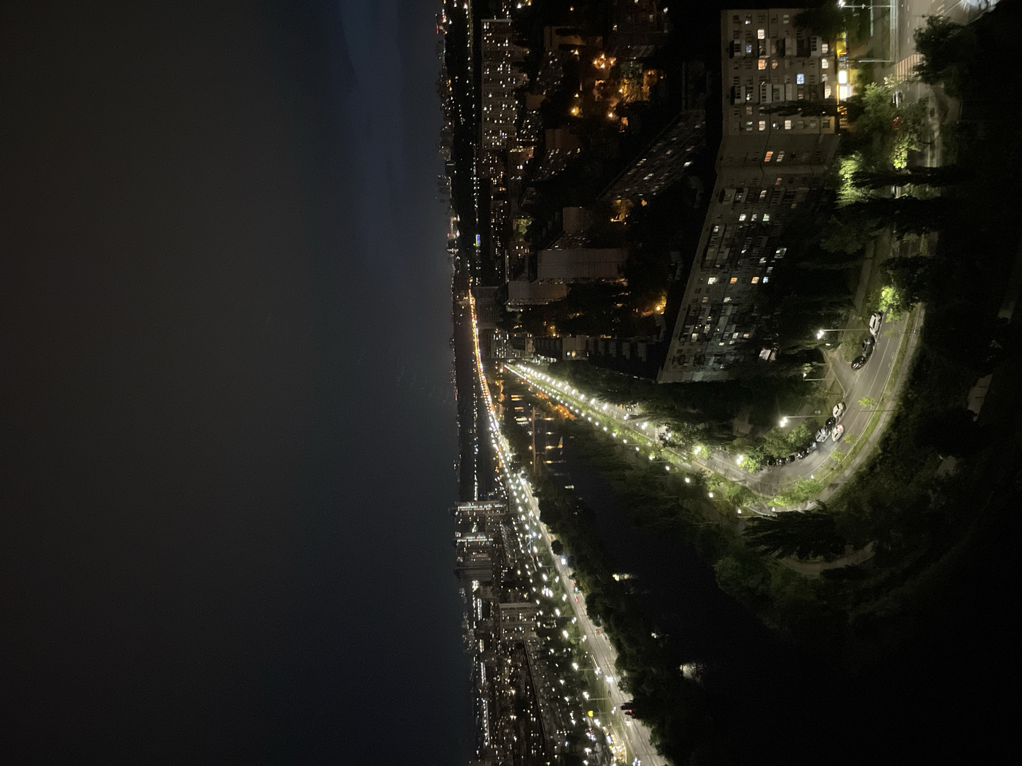 This is an aerial nighttime photograph of a city in Kyiv, Ukraine, likely taken from a high vantage point. The scene is dominated by the glowing lights of a large urban area under a dark, cloudy sky.
In the foreground and midground, a winding road is illuminated by numerous streetlights, casting a bright, greenish-white glow. Cars are visible on the road, with some appearing as streaks of light indicating motion. Adjacent to the road, a dark body of water, possibly a river or canal, reflects some of the surrounding lights. Lush green trees line the banks of the water and the roadside, appearing dark silhouettes against the artificial illumination.
Further into the midground, numerous apartment buildings are visible. Their windows are lit with a warm yellow and orange light, suggesting occupancy. Some of these buildings are tall and modern, while others appear older and more uniform in design. Construction cranes are visible near some of the taller structures, indicating ongoing development.
In the background, the city extends as far as the eye can see, with a dense concentration of lights suggesting a vast urban landscape. A brightly lit bridge or highway stretches across the horizon, further emphasizing the extensive infrastructure. The overall impression is of a vibrant city alive with activity at night, despite the darkness of the sky. The image captures the interplay of artificial light and natural darkness, highlighting the urban environment.
