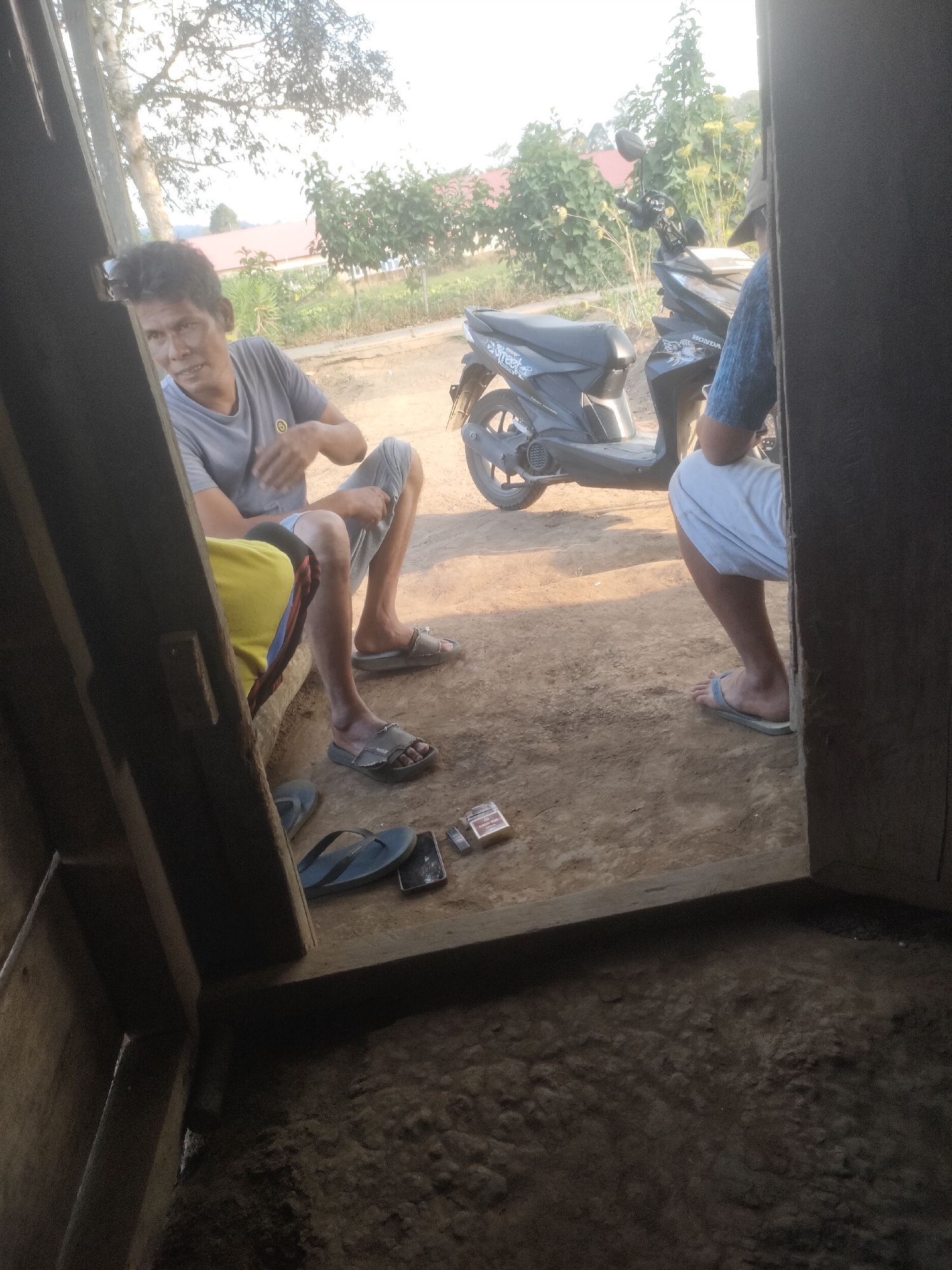 The image depicts an outdoor scene in Padang Dalom, Indonesia, showing three individuals, two seated and one standing. A man in a grey t-shirt and shorts is seated on the ground, leaning against a wooden structure. To his right, another person in a blue shirt and white shorts is also seated. A third individual, mostly obscured by a wooden doorway, is standing near a dark-colored motorcycle. The ground is dirt and pebbles. Several pairs of flip-flops, a smartphone, and a pack of cigarettes are scattered on the ground. In the background, there are trees and buildings. The lighting suggests it is daytime, possibly late afternoon, with sunlight casting shadows.