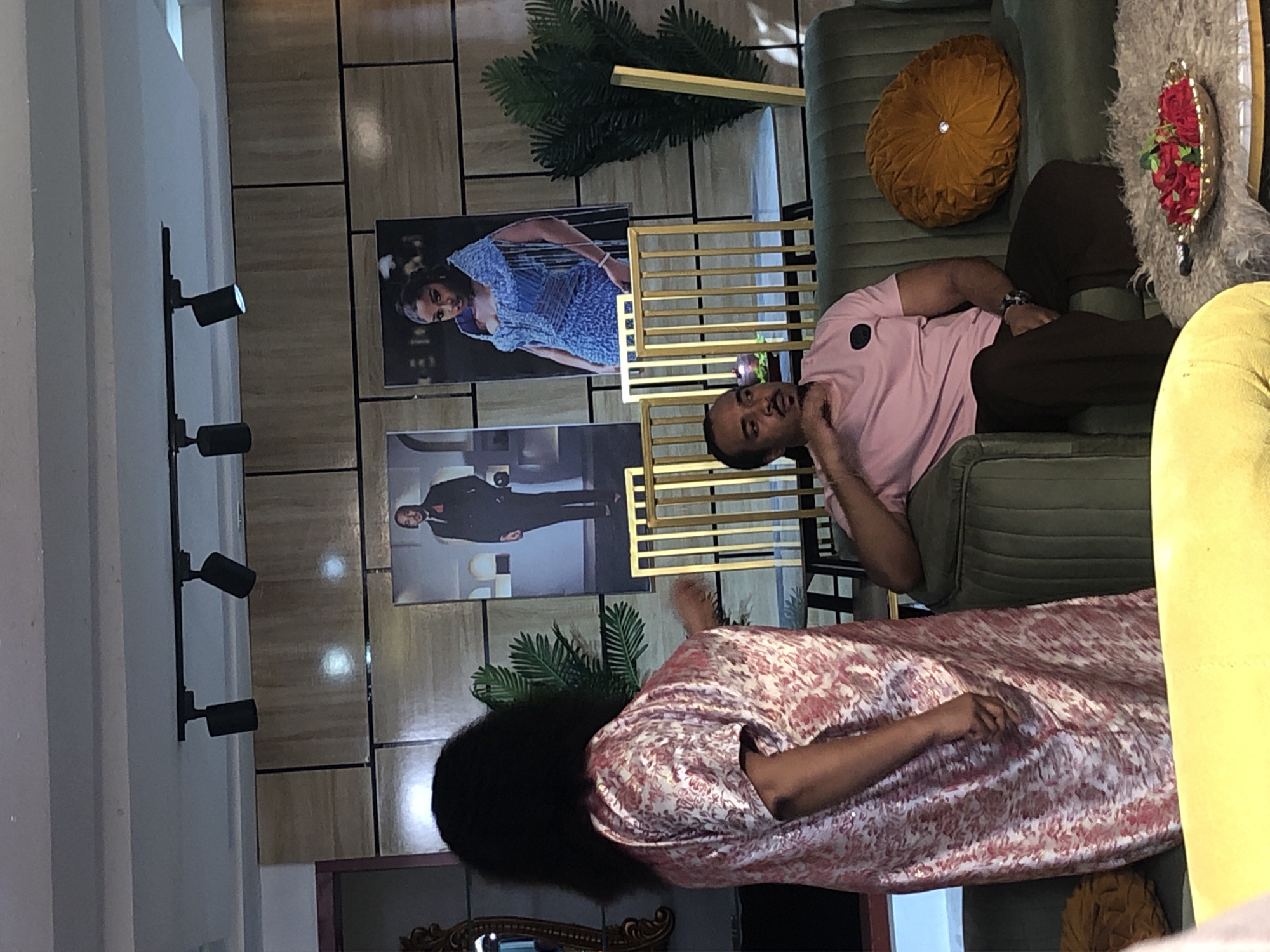 A man is sitting on a green couch in what appears to be an indoor studio setting. He is wearing a pink t-shirt and brown pants. Next to him is a woman, partly visible, wearing a red and white patterned dress. On the wall behind them are two pictures, one of a woman in a blue dress and another of a man in a suit. There is a gold frame and some plants decorating the wall. The lighting appears to come from the left side of the frame. The exact location and city are not discernible from the image.