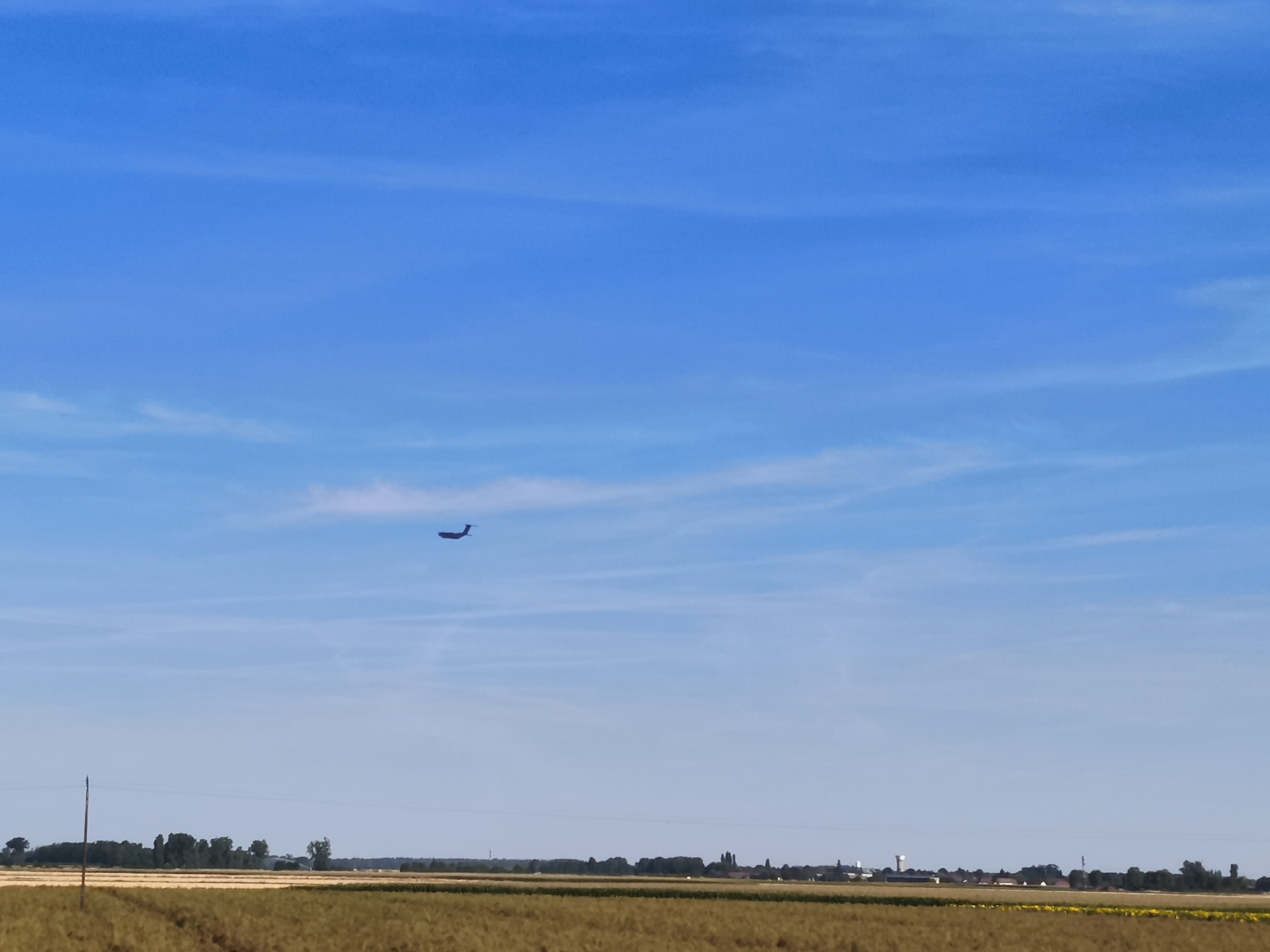 The image captures a wide-angle view of a rural landscape under a clear blue sky with sparse wispy clouds. In the upper portion of the sky, a dark silhouette of an airplane is visible, flying from left to right. The horizon line is low in the frame, dividing the sky from the land. Below the sky, a strip of trees forms a dark line across the mid-ground, with a few scattered buildings and structures visible behind them. The foreground is dominated by a field of dry, golden-brown vegetation, likely crops or hay, with some tire tracks visible on its surface. A solitary wooden utility pole stands on the left side of the frame, rising from the field. The overall impression is one of a sunny, tranquil day in the countryside. The image does not provide specific details that would allow for pinpointing the exact location within Peseux, France, but it depicts a typical rural setting found in such areas. There are no people visible in the image.